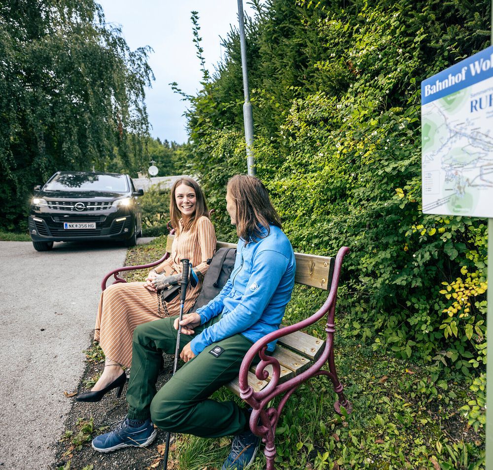 Ein schwarzer Kleinbus, der RUF Bus, holt gerade eine Frau in einem schönen orangen Kleid und einen Wanderer ab. 