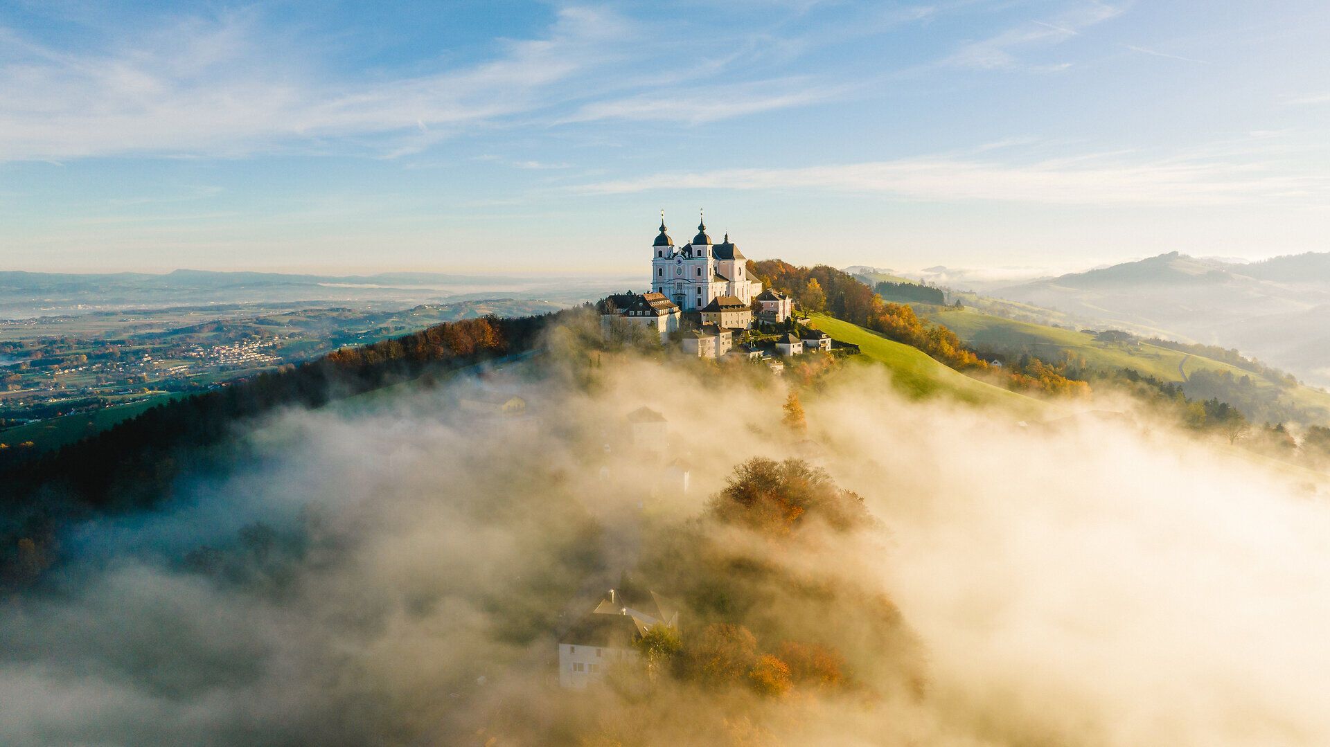 Drohnenaufnahme der Basilika Sonntagberg im Mostviertel, die auf einem Hügel über einer von Nebel durchzogenen Landschaft mit sanften Hügeln und Herbstfärbung thront.