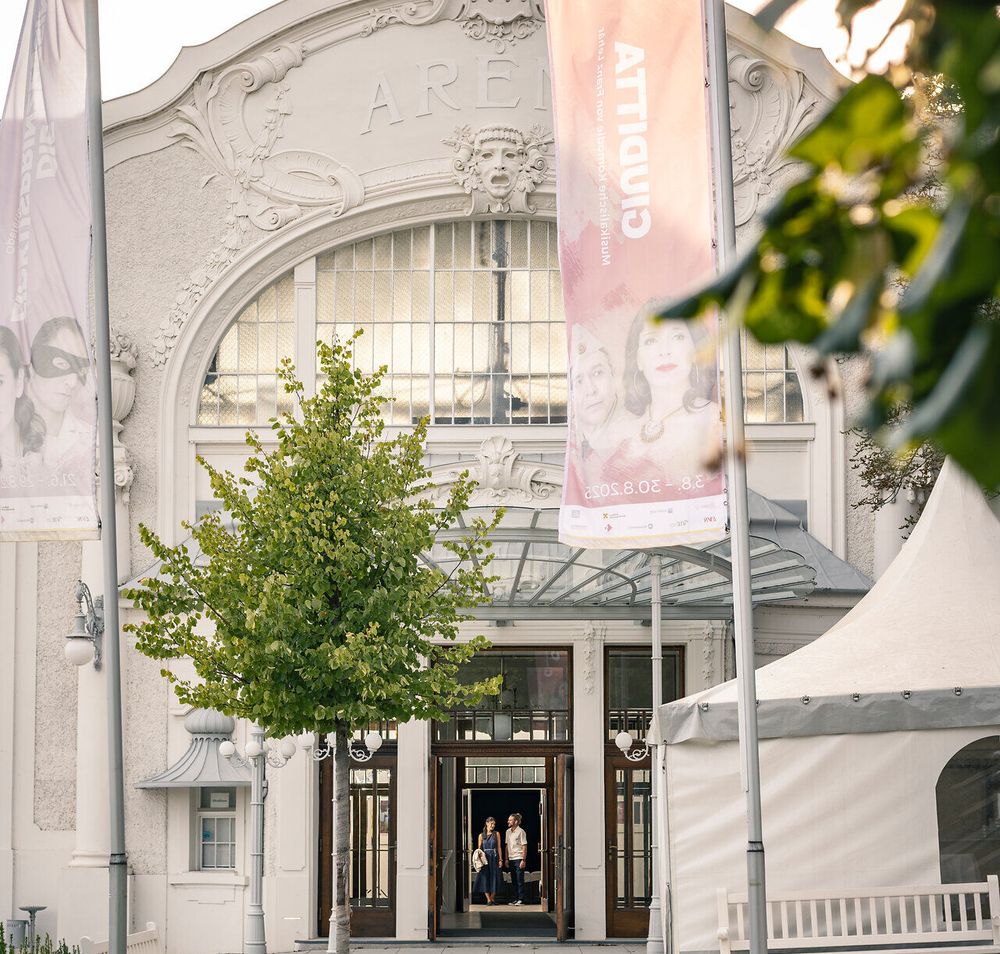 Ein Paar verlässt die historische Sommerarena Baden, ein beeindruckendes Bauwerk. Die Sonne über Baden bei Wien taucht die Szene in warmes Licht, während die beiden das Freigelände betreten und der kulturellen Atmosphäre nachspüren.