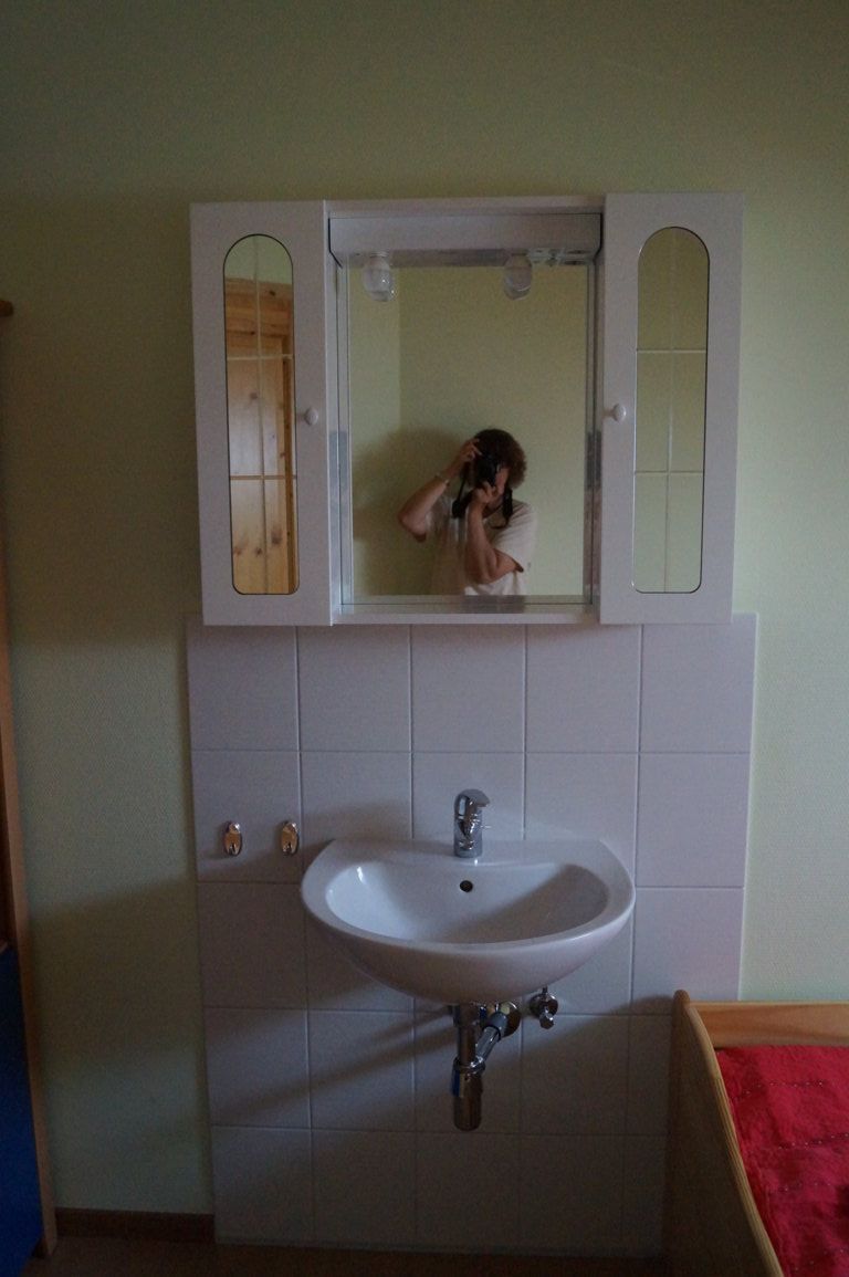 Ein Waschbecken mit Spiegel und Schrank in einem Zimmer. Eine Person fotografiert sich im Spiegel.