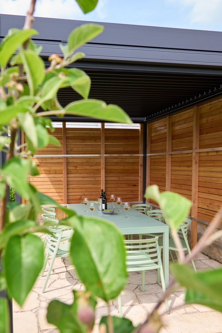Überdachte Terrasse mit Tisch und Stühlen, umgeben von Holzverkleidung und Pflanzen im Vordergrund.