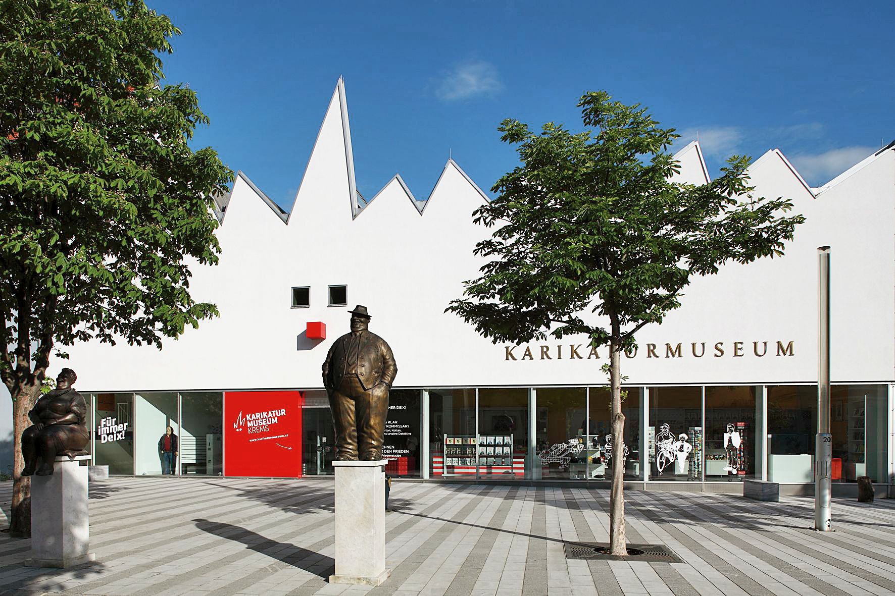 Außenansicht des Karikaturmuseums Krems mit Statuen und Bäumen.