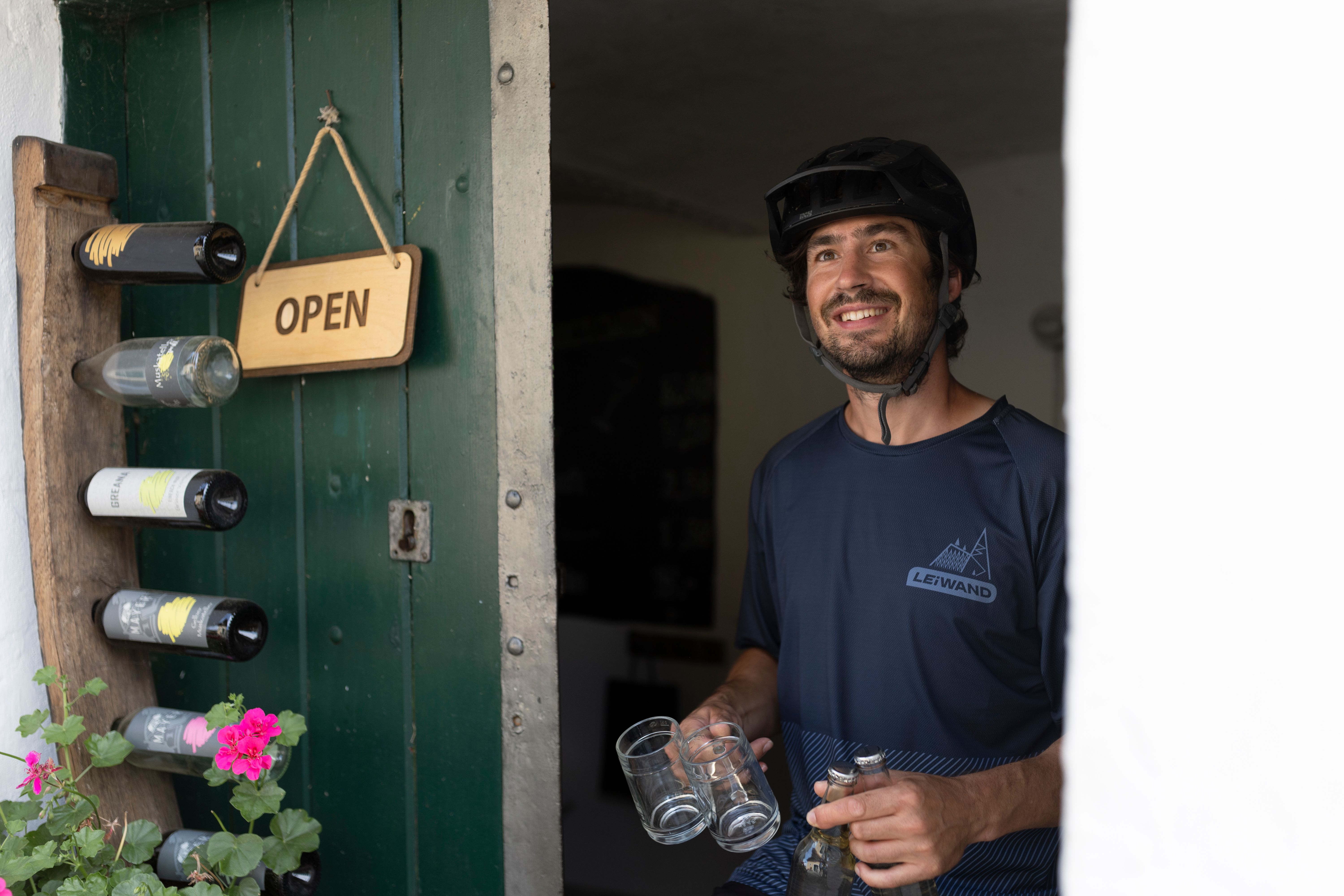 Ein Mann mit Fahrradhelm steht lächelnd in einer offenen Tür, hält Gläser und Flaschen. An der Tür hängt ein "Open"-Schild.