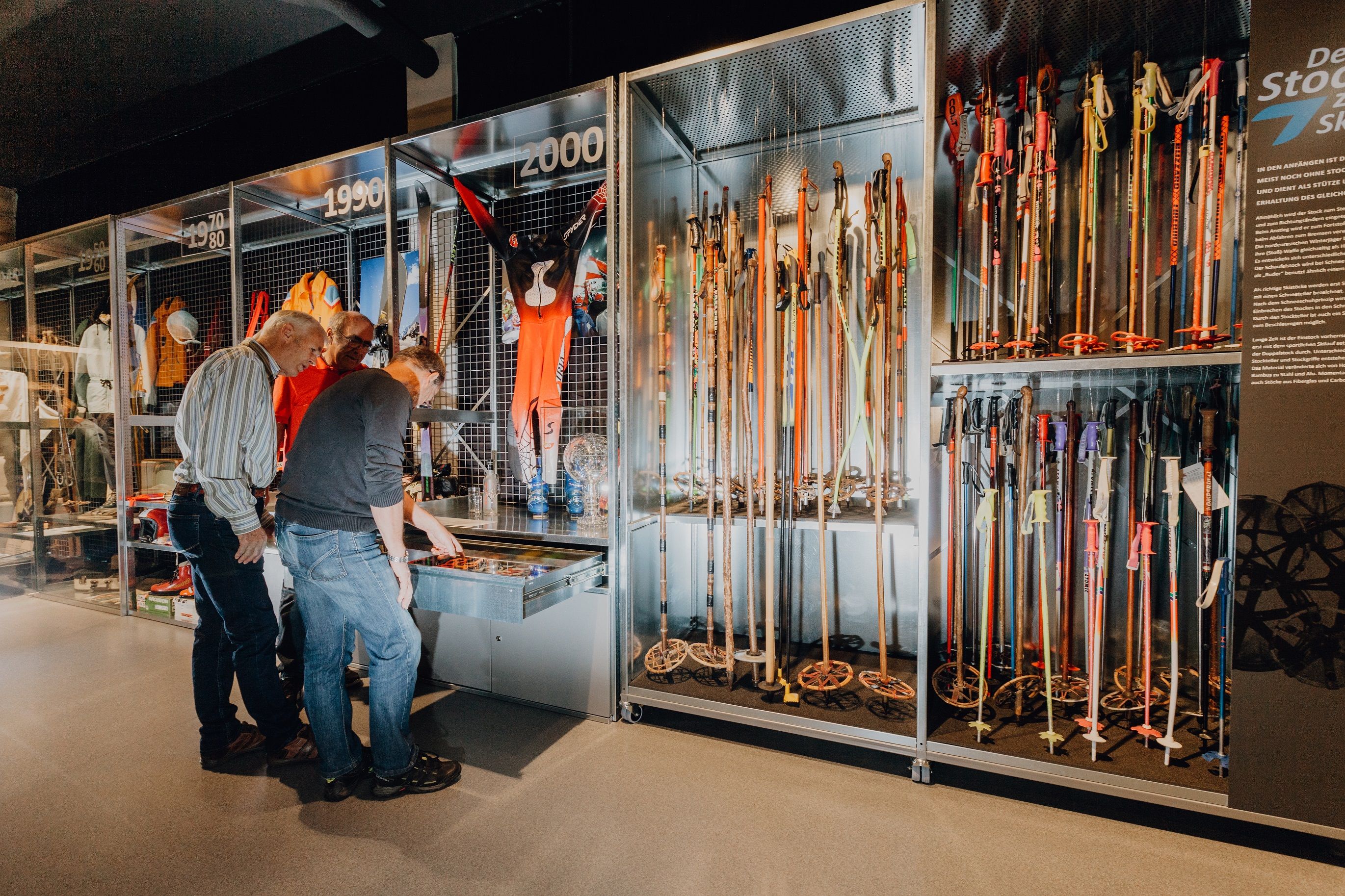 Drei Männer betrachten im Wintersportmuseum eine Ausstellung mit historischen Skistöcken und Wintersportausrüstung in Vitrinen.