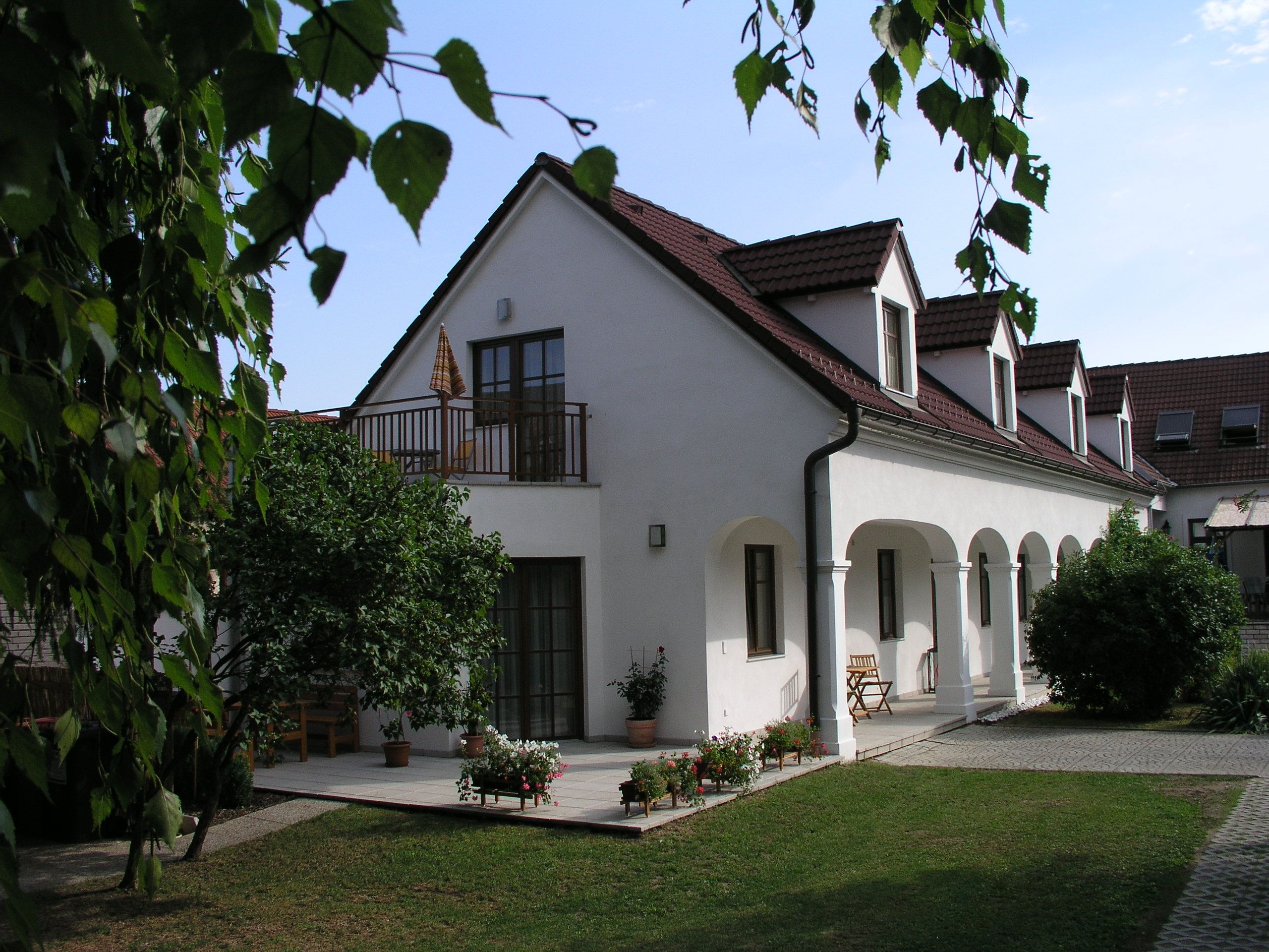 Ein weißes Gebäude mit roten Dachziegeln und Arkaden, umgeben von einem Garten mit Bäumen und Blumen.