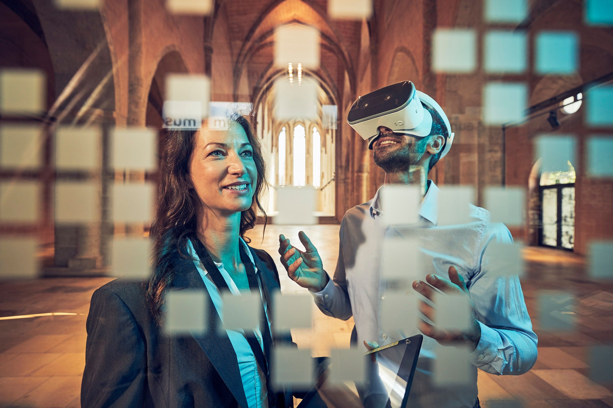 Zwei Personen in einem historischen Gebäude, eine trägt eine VR-Brille, die andere lächelt. Im Vordergrund sind unscharfe Quadrate zu sehen.