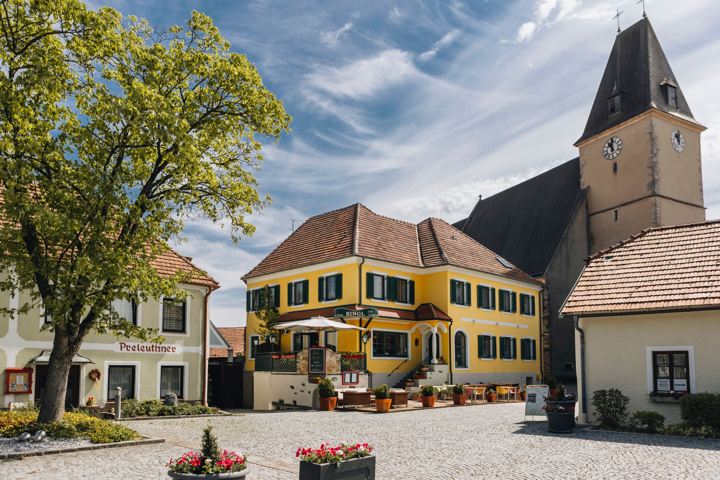 Idyllischer Dorfplatz mit Gasthaus und Terrasse und ein Kirchturm im Hintergrund.