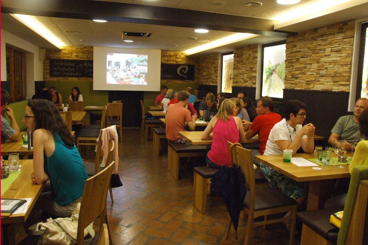 Menschen sitzen in einem gemütlichen Raum an Holztischen, während eine Präsentation auf einer Leinwand gezeigt wird.
