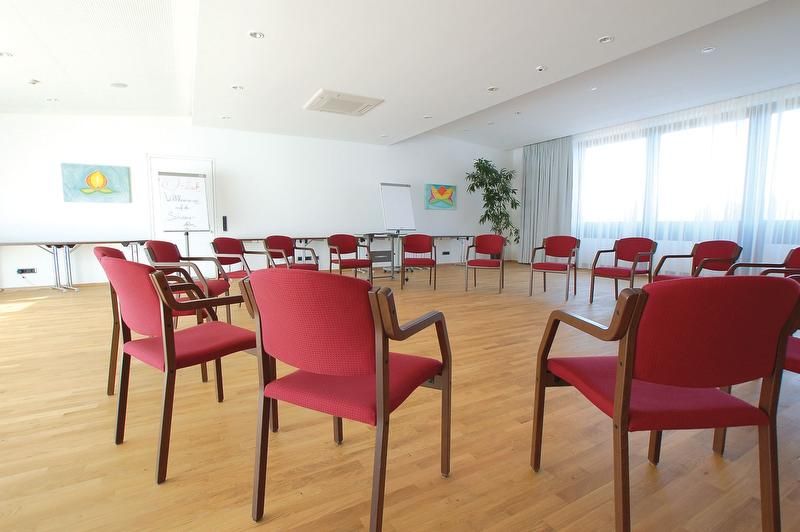 Ein heller Seminarraum mit roten Stühlen im Kreis, Holzboden und großen Fenstern.