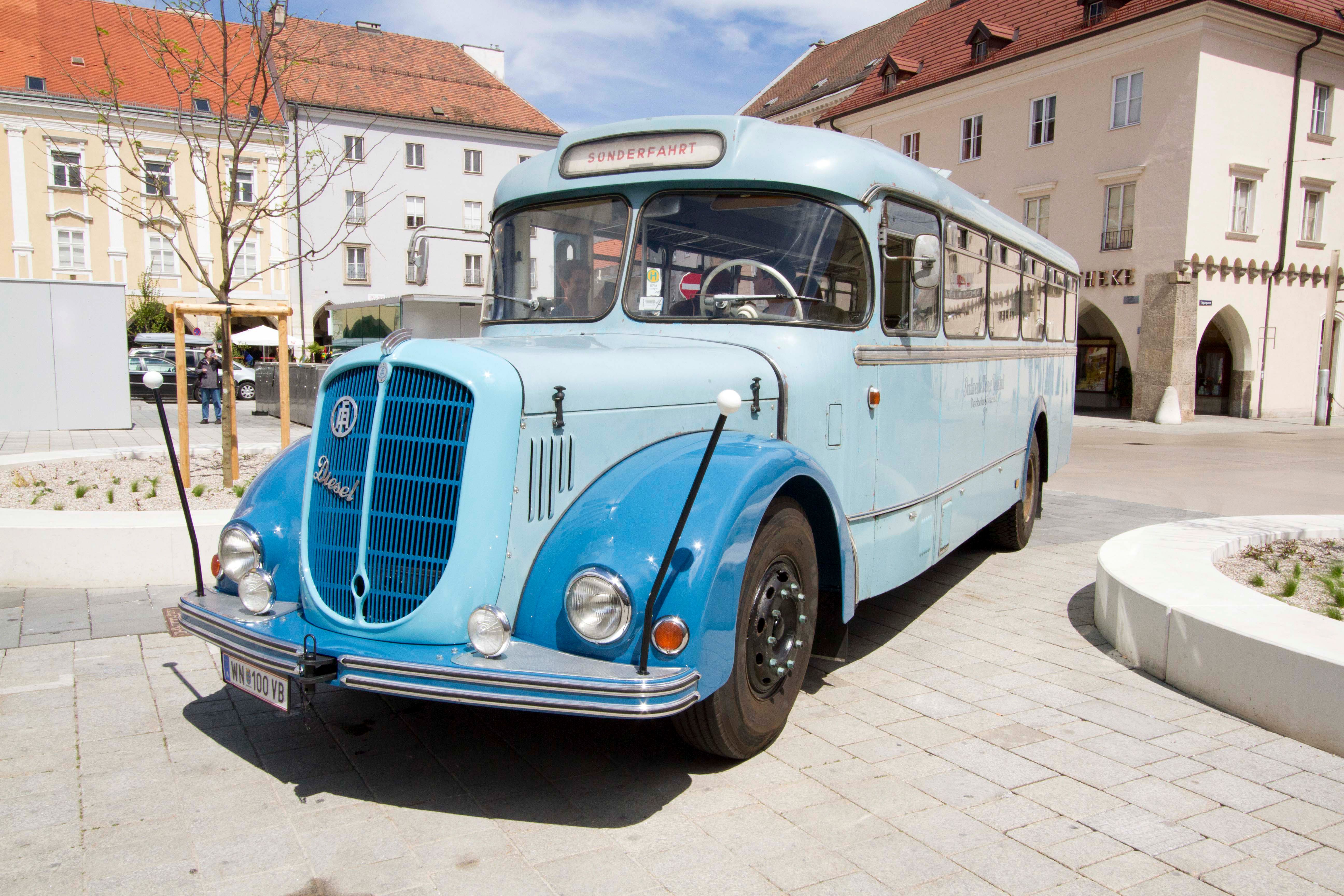 Ein blauer Oldtimer-Bus steht auf einem gepflasterten Platz vor historischen Gebäuden.