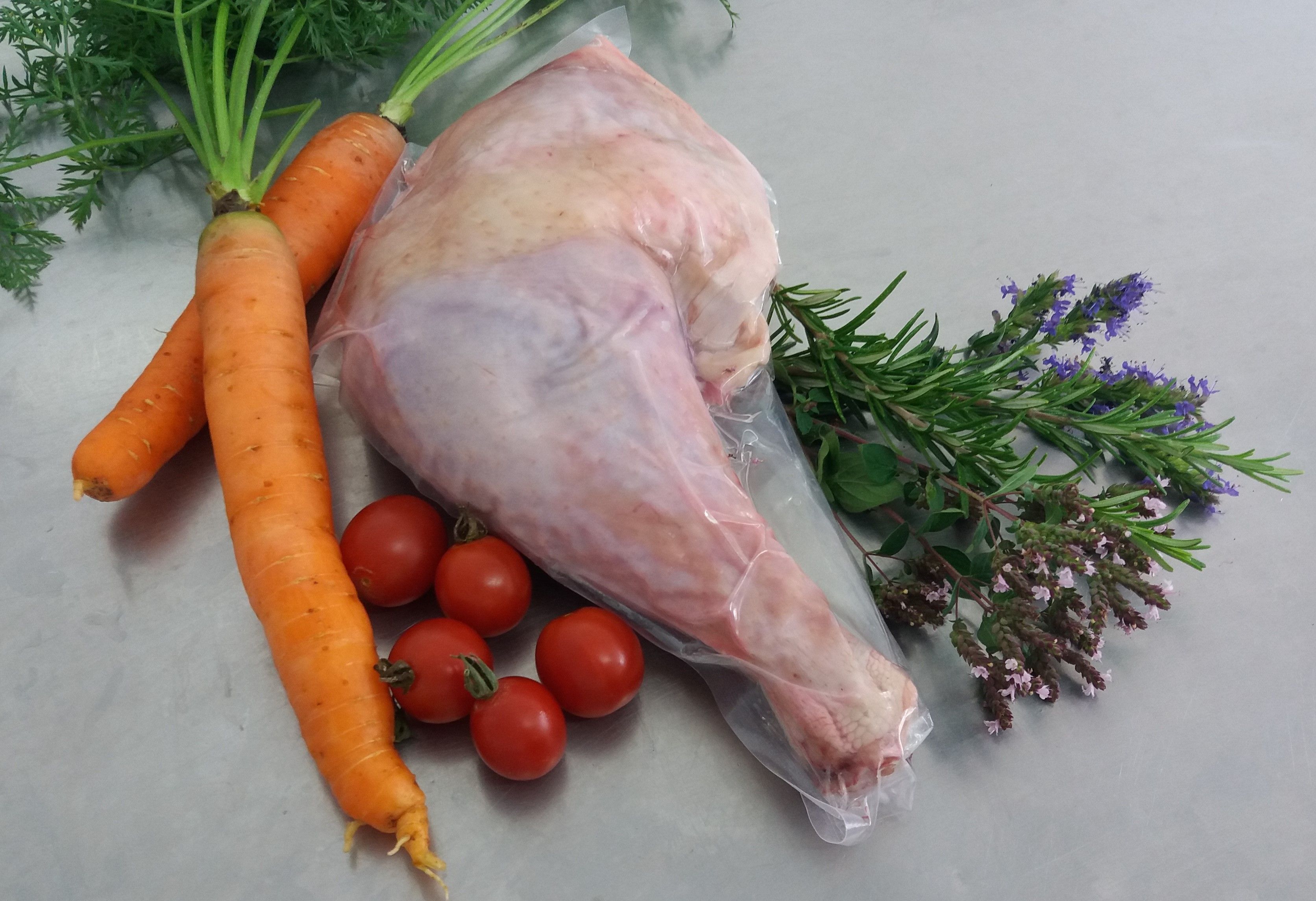 Eine Putenkeule in Plastik verpackt mit Karotten, Tomaten und Kräuten auf einem hellgrauen Hintergrund.