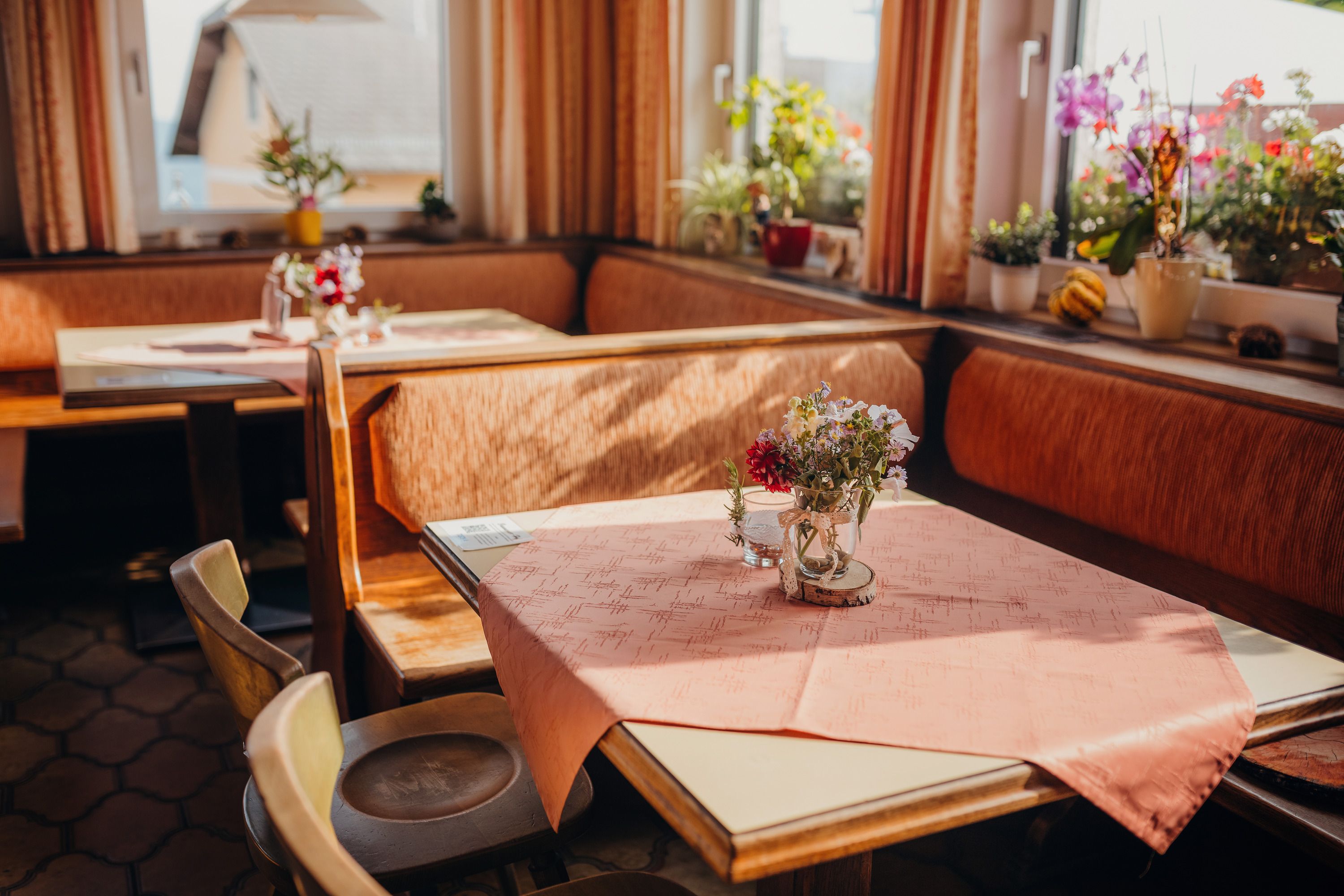 Gemütliche Stube mit Holzmöbeln, Tischdecken und Blumen auf den Tischen, umgeben von Fenstern mit Pflanzen.