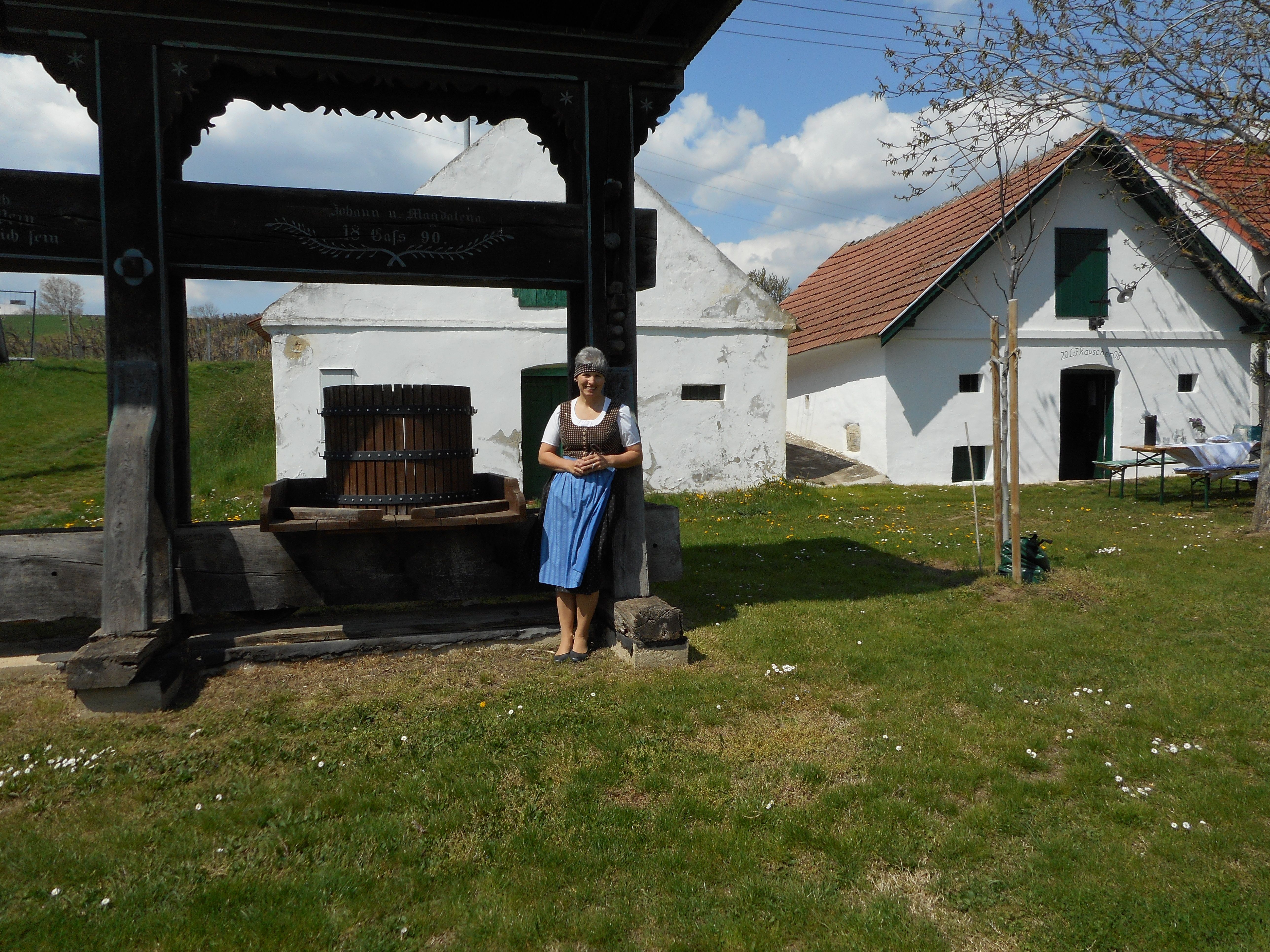 Eine Frau in traditioneller Kleidung steht neben einer alten Weinpresse vor einem historischen Gebäude mit weißen Wänden und roten Dächern.