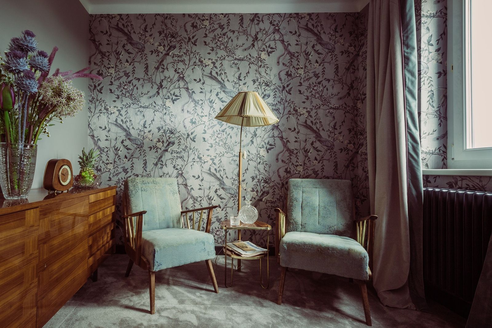 Ein stilvoll eingerichtetes Zimmer mit zwei blauen Sesseln, einer Stehlampe und einer Kommode mit Blumen und einer Uhr.