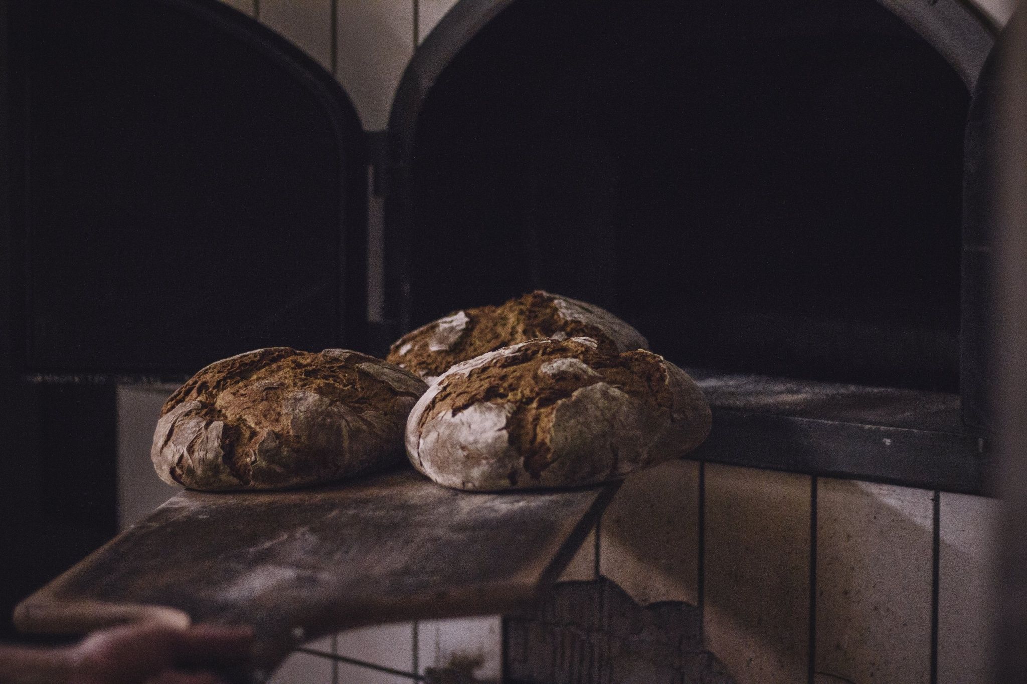 Drei Brote auf einem Holzschieber vor einem Backofen.