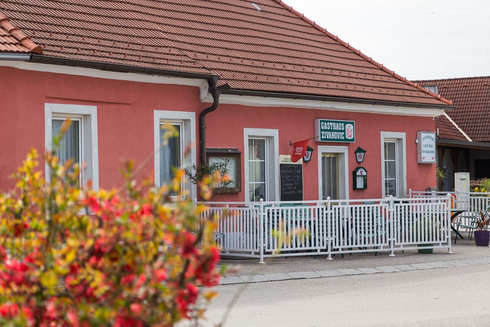 Ein rosa Gebäude mit der Aufschrift 'Gasthaus Zivanovic', umgeben von einem weißen Zaun und Blumen im Vordergrund.
