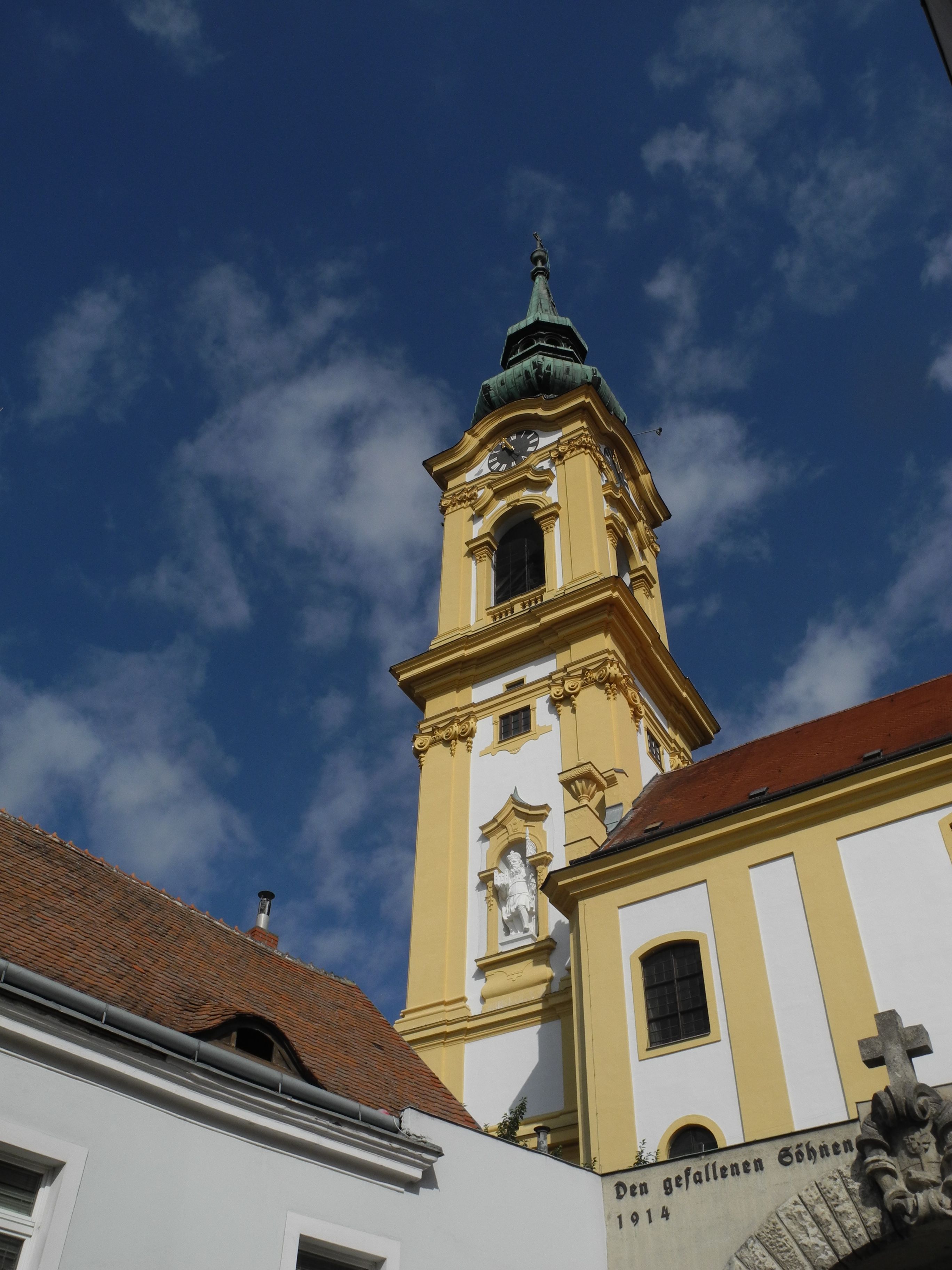 Gelber Kirchturm der Stadtpfarrkirche Stockerau vor blauem Himmel.