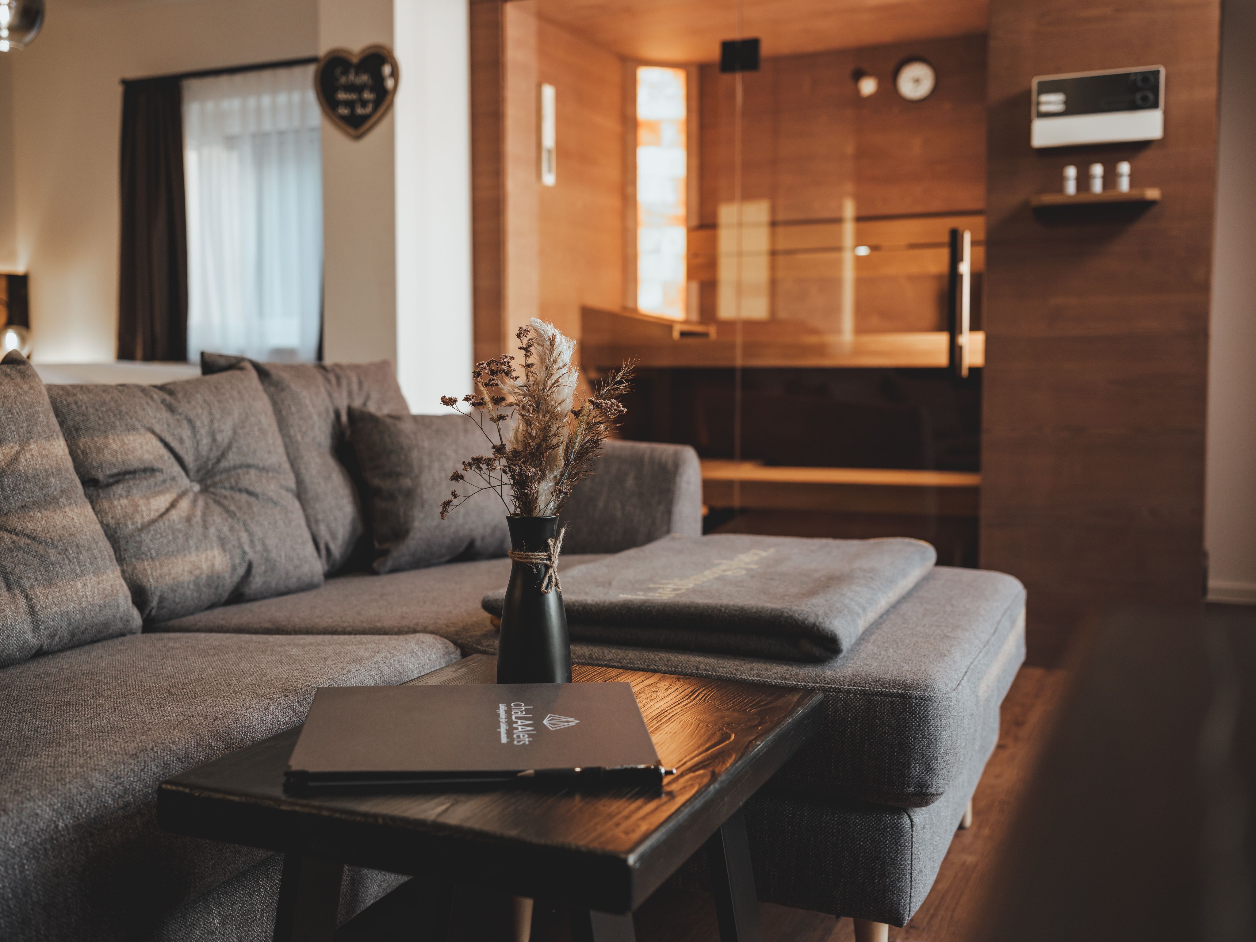 Gemütliches Wohnzimmer mit grauem Sofa, Holztisch und Sauna im Hintergrund.