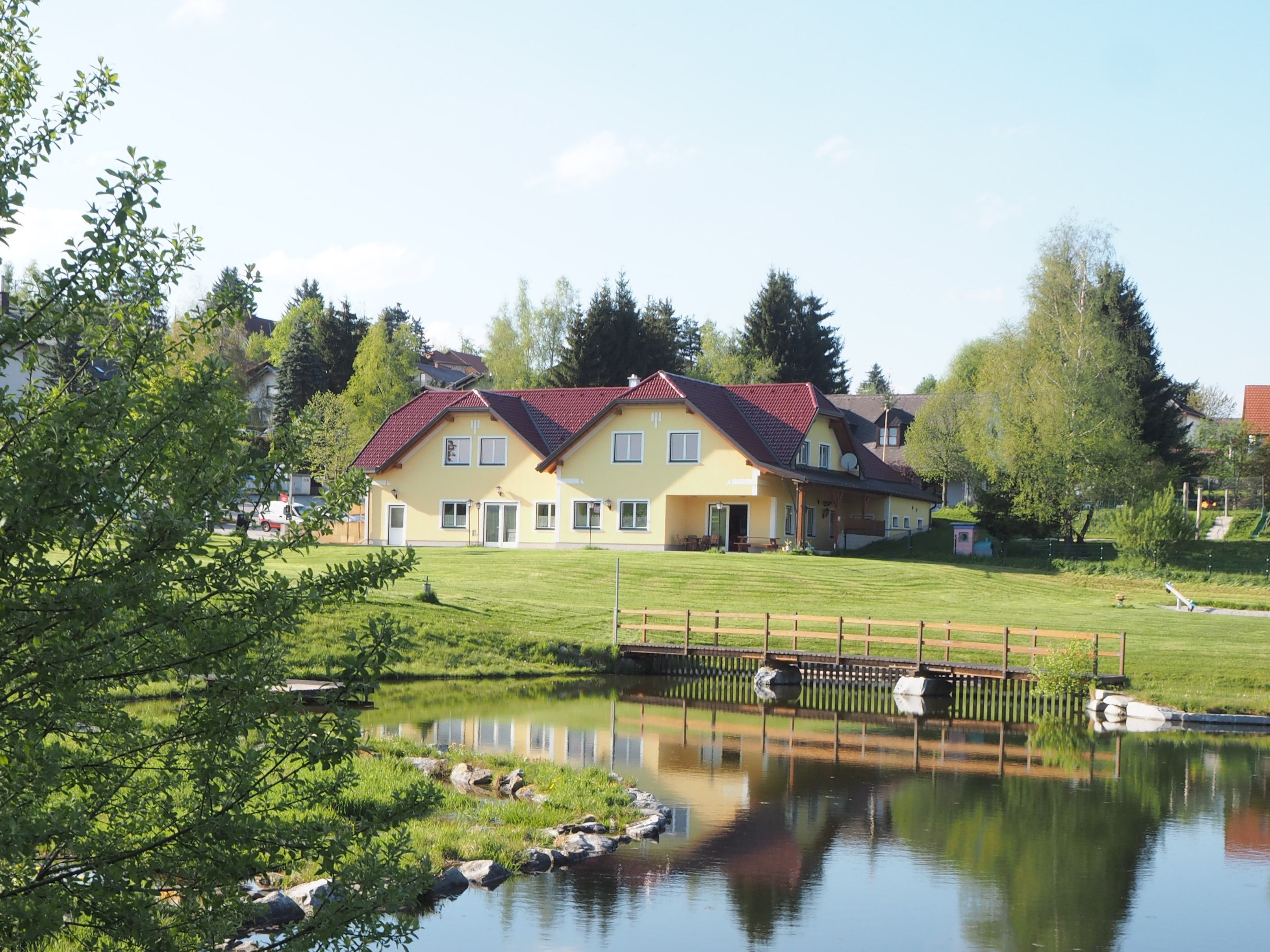 Gelbes Gebäude mit rotem Dach an einem Teich, umgeben von Bäumen und Rasen.