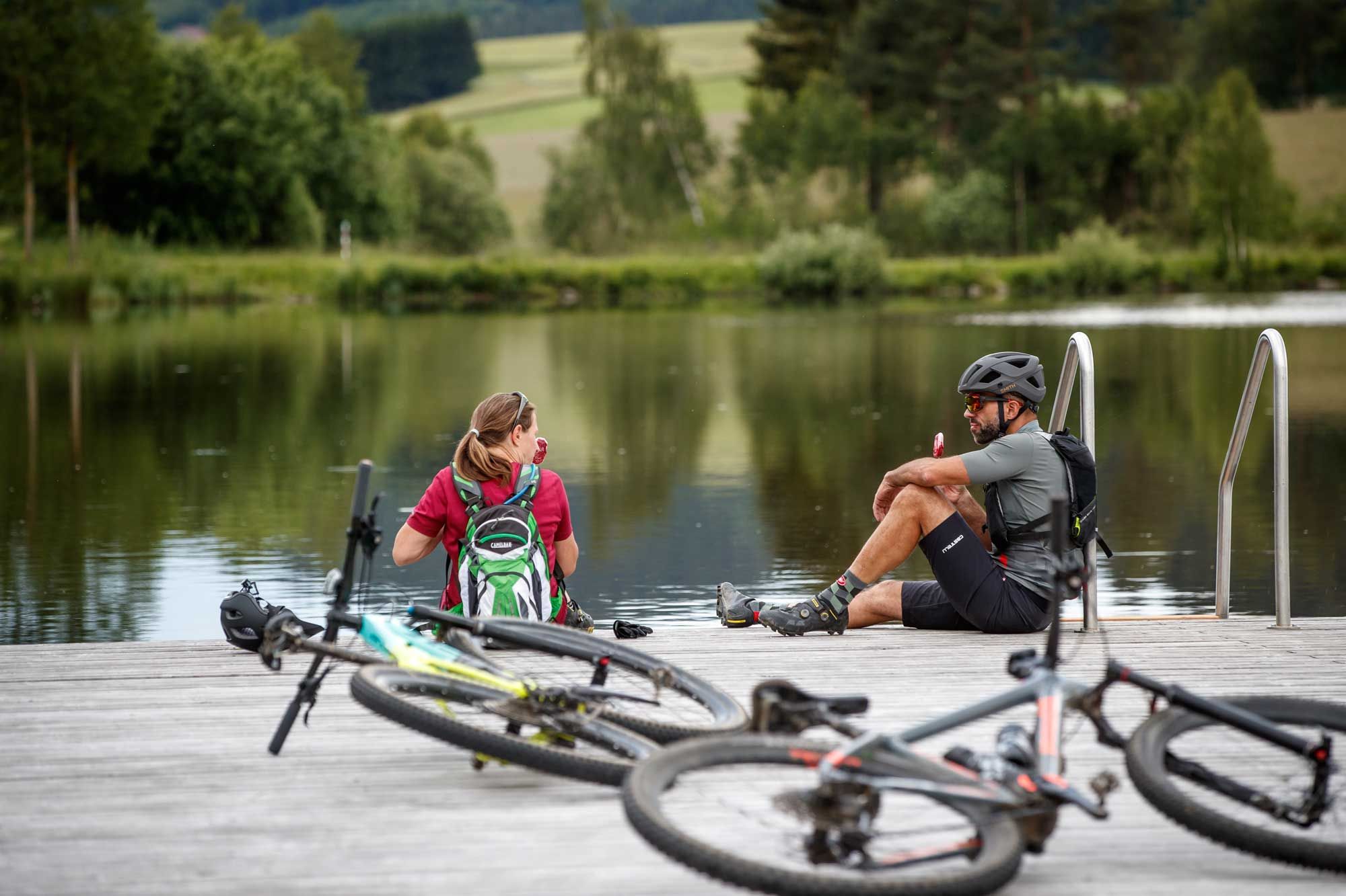 Zwei Radfahrer sitzen auf einem Steg am Frauenwieserteich, umgeben von Fahrrädern und Natur.