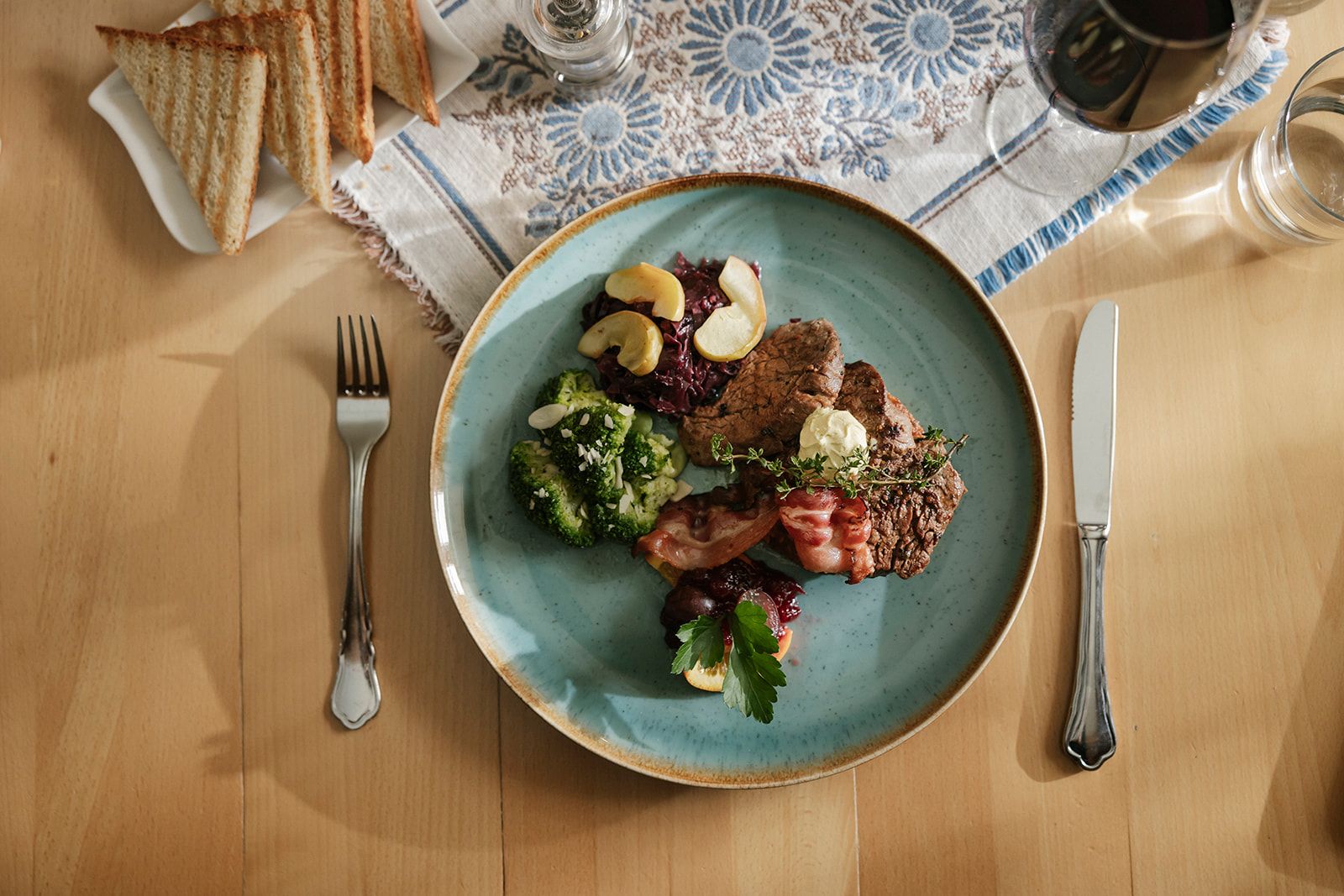 Ein Teller mit Steak, Brokkoli, Speck und Rotkohl auf einem Tisch mit Besteck und Toast.