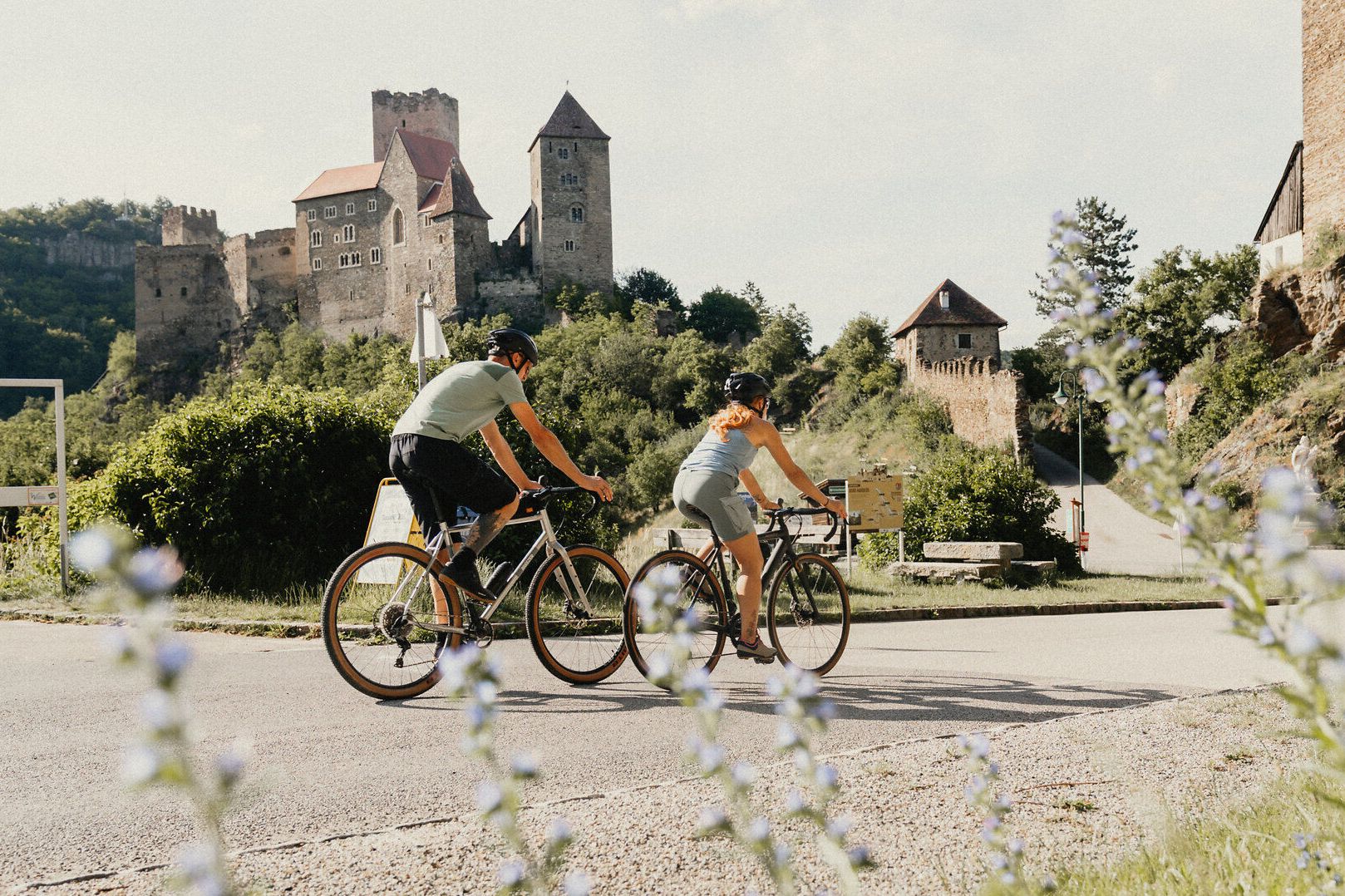 Zwei Radfahrer fahren entlang eine Straße, hinter ihnen die Burg Hardegg, im Vordergrund Blumen