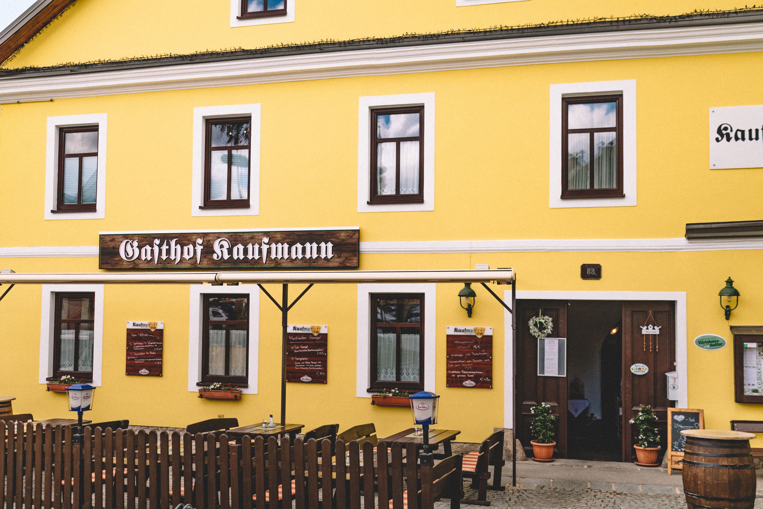 Gelbes Gebäude mit der Aufschrift 'Gasthof Kaufmann', Holztische und Stühle im Freien.