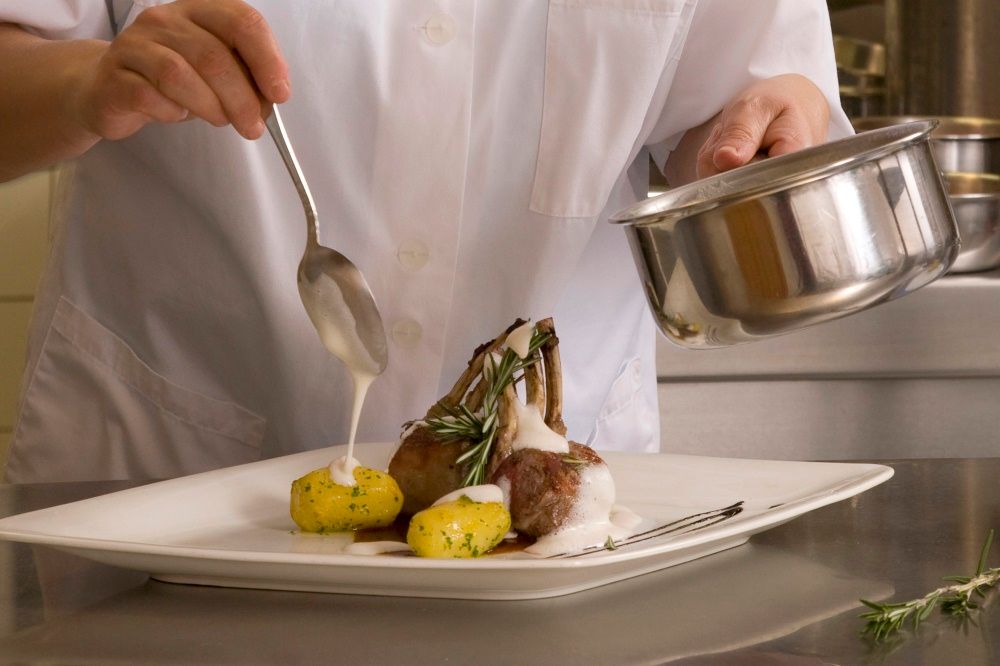 Ein Koch richtet ein Gericht mit Lammkoteletts und Kartoffeln auf einem Teller an.