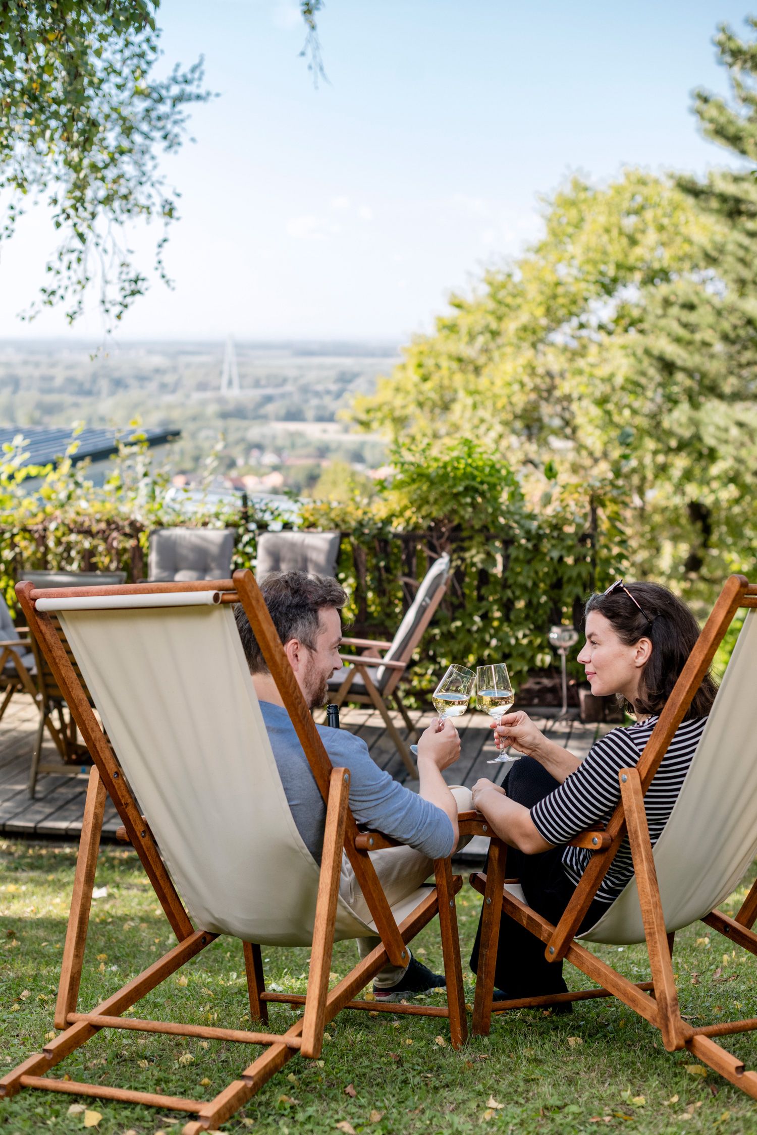 Ein Paar sitzt in Liegestühlen im Garten und stößt mit Weingläsern an, mit Blick auf eine grüne Landschaft.