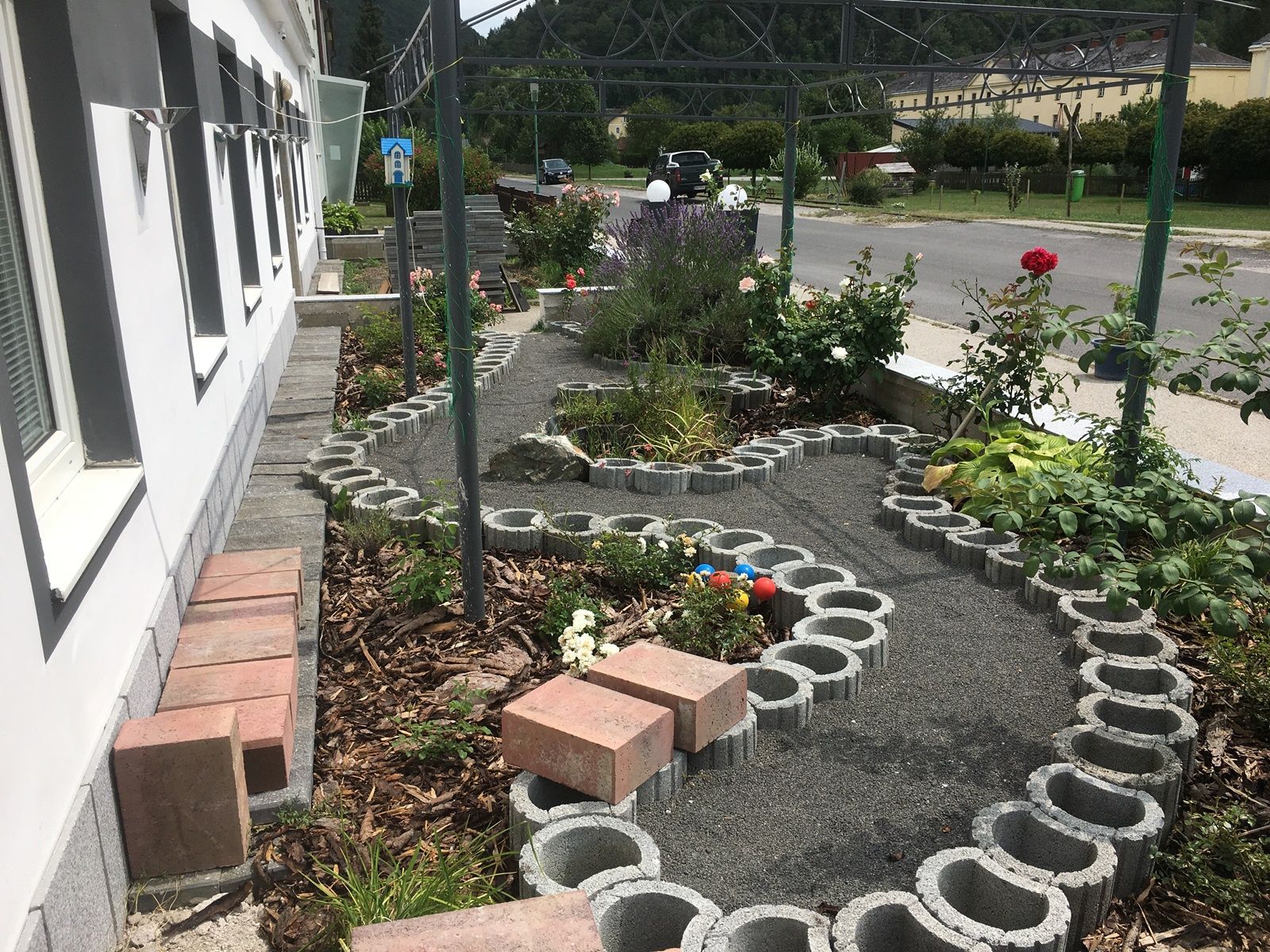 Ein kleiner Garten mit Rosen und einem geschwungenen Weg aus Betonsteinen neben einem Gebäude.