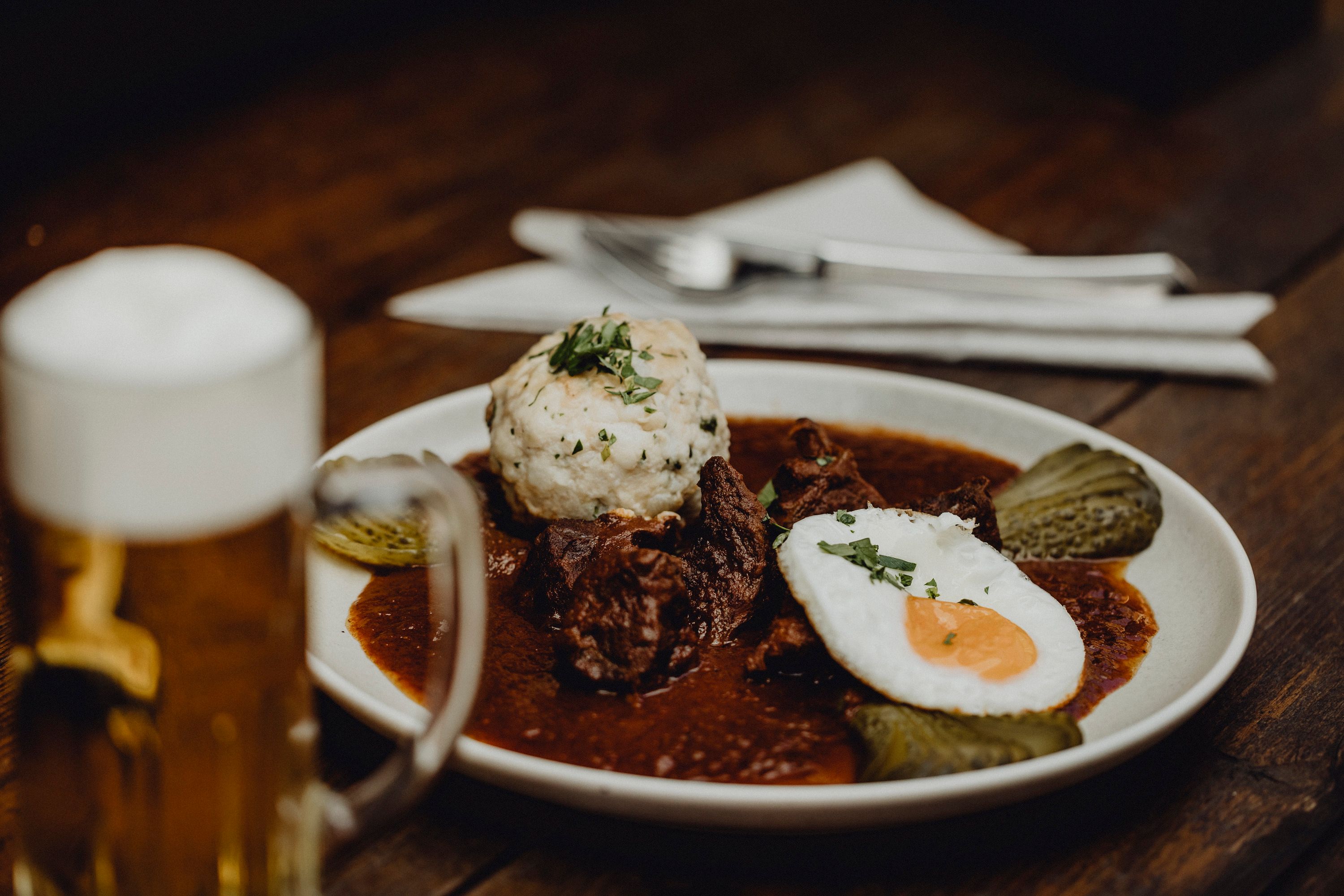Ein Teller mit Gulasch, Knödel, Spiegelei und Gurken, daneben ein Bierkrug auf einem Holztisch.