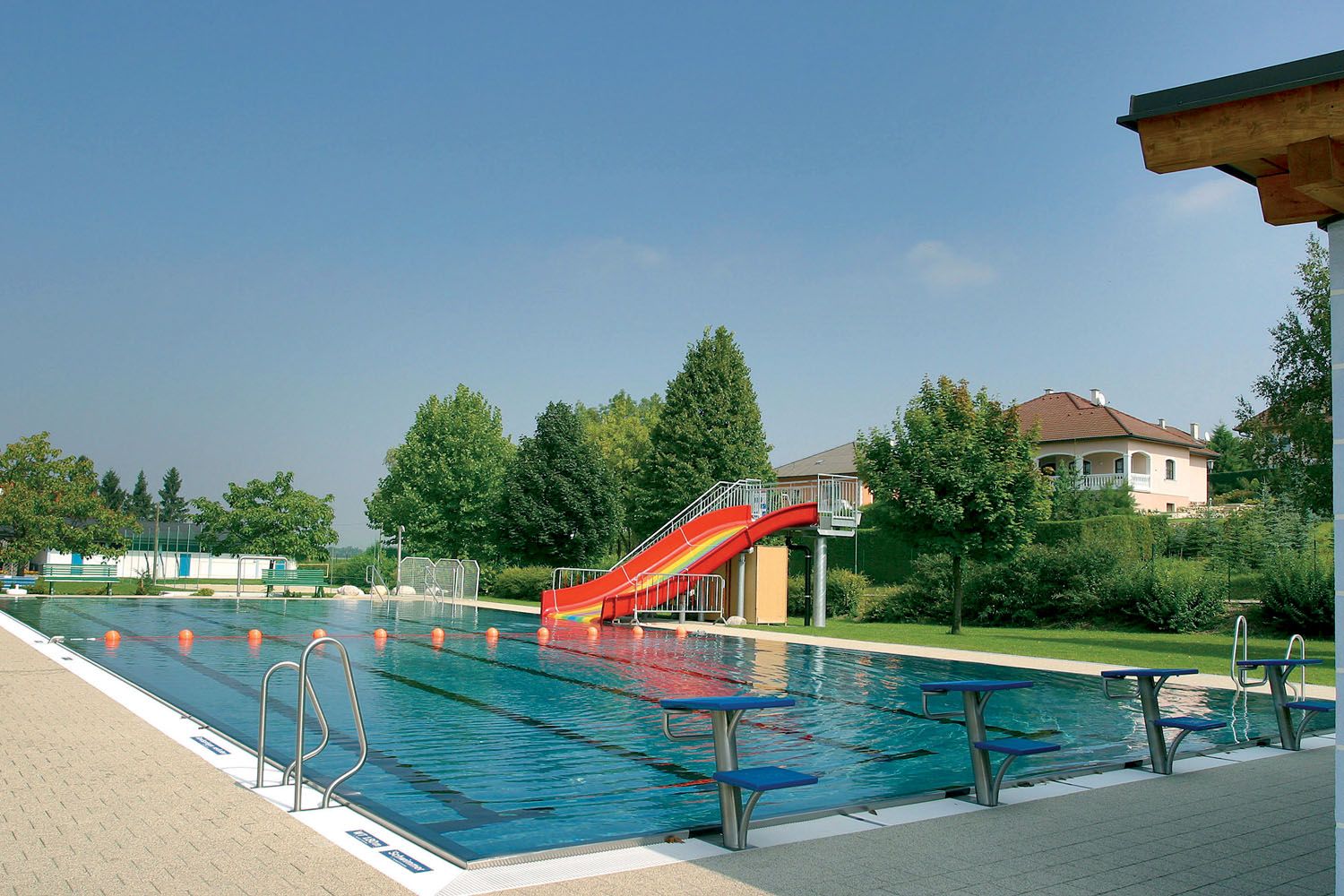Ein Freibad mit einem Schwimmbecken, einer roten Rutsche und Sprungbrettern. Im Hintergrund sind Bäume und ein Haus zu sehen.