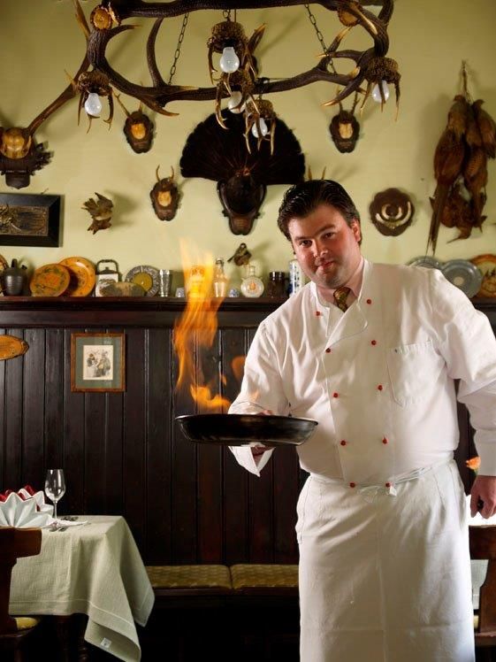 Ein Koch in weißer Uniform flambiert eine Speise in einer rustikalen Küche mit Jagdtrophäen an der Wand.