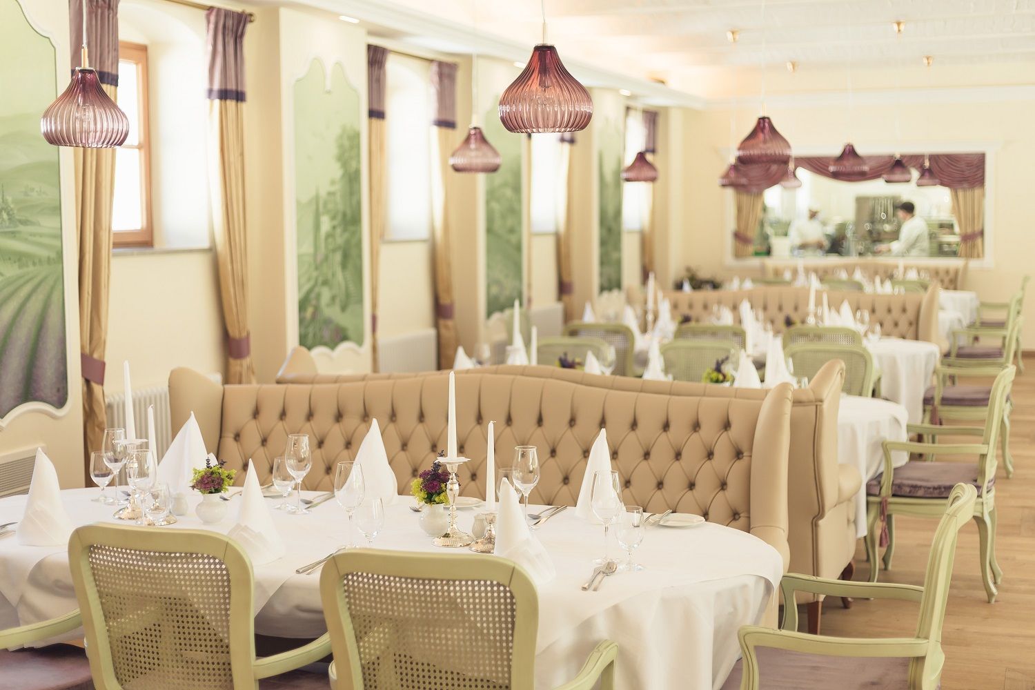 Elegantes Restaurant mit gedeckten Tischen, weißen Tischdecken und Kerzenleuchtern, beige Polsterbänke und grüne Stühle, dekorative Wandmalereien.