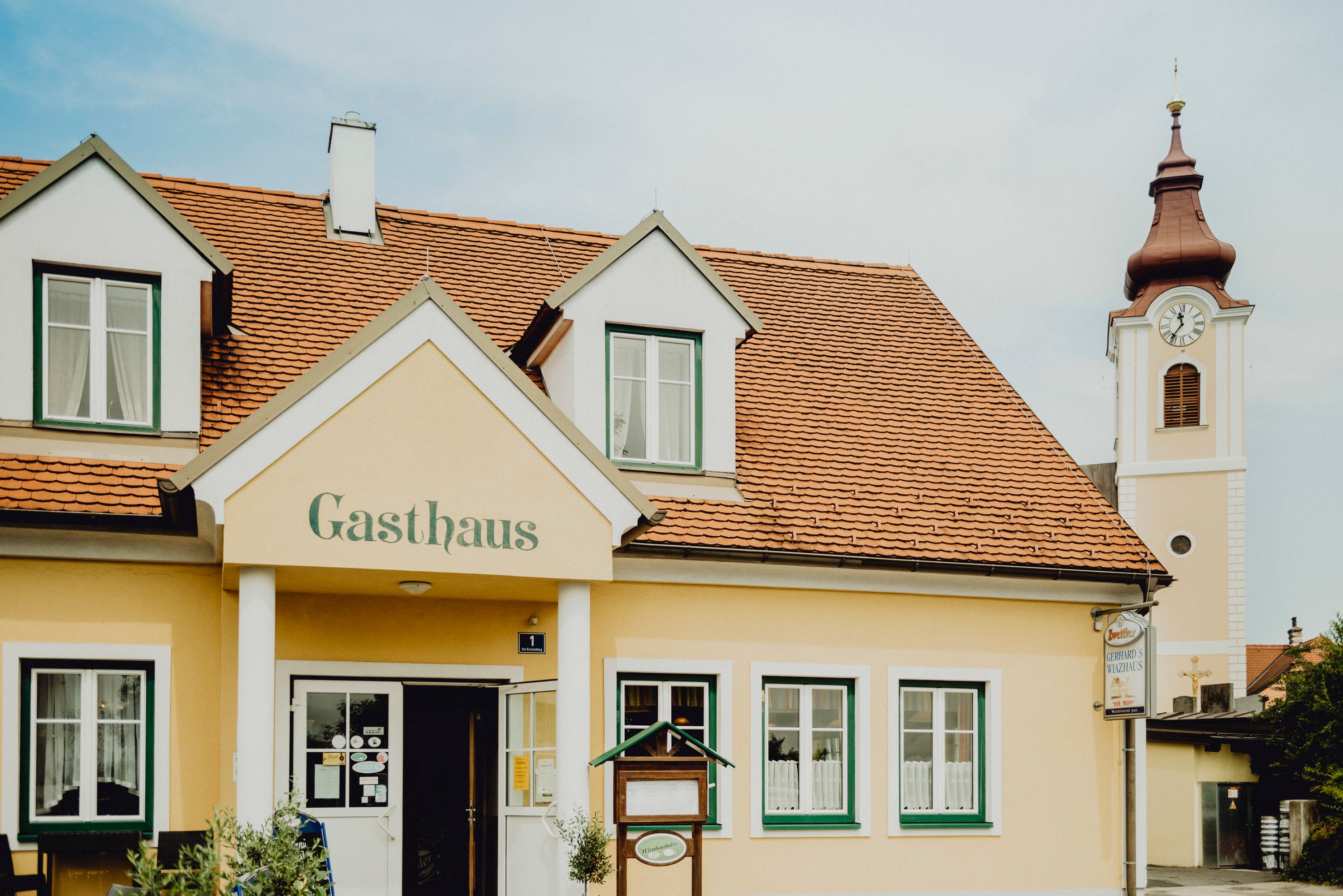 Ein gelbes Gasthaus mit roten Dachziegeln und einem Kirchturm im Hintergrund.