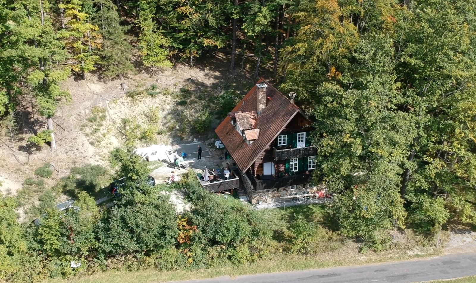 Luftaufnahme eines Hauses im Wald mit rotem Dach und grünen Fensterläden, umgeben von Bäumen.