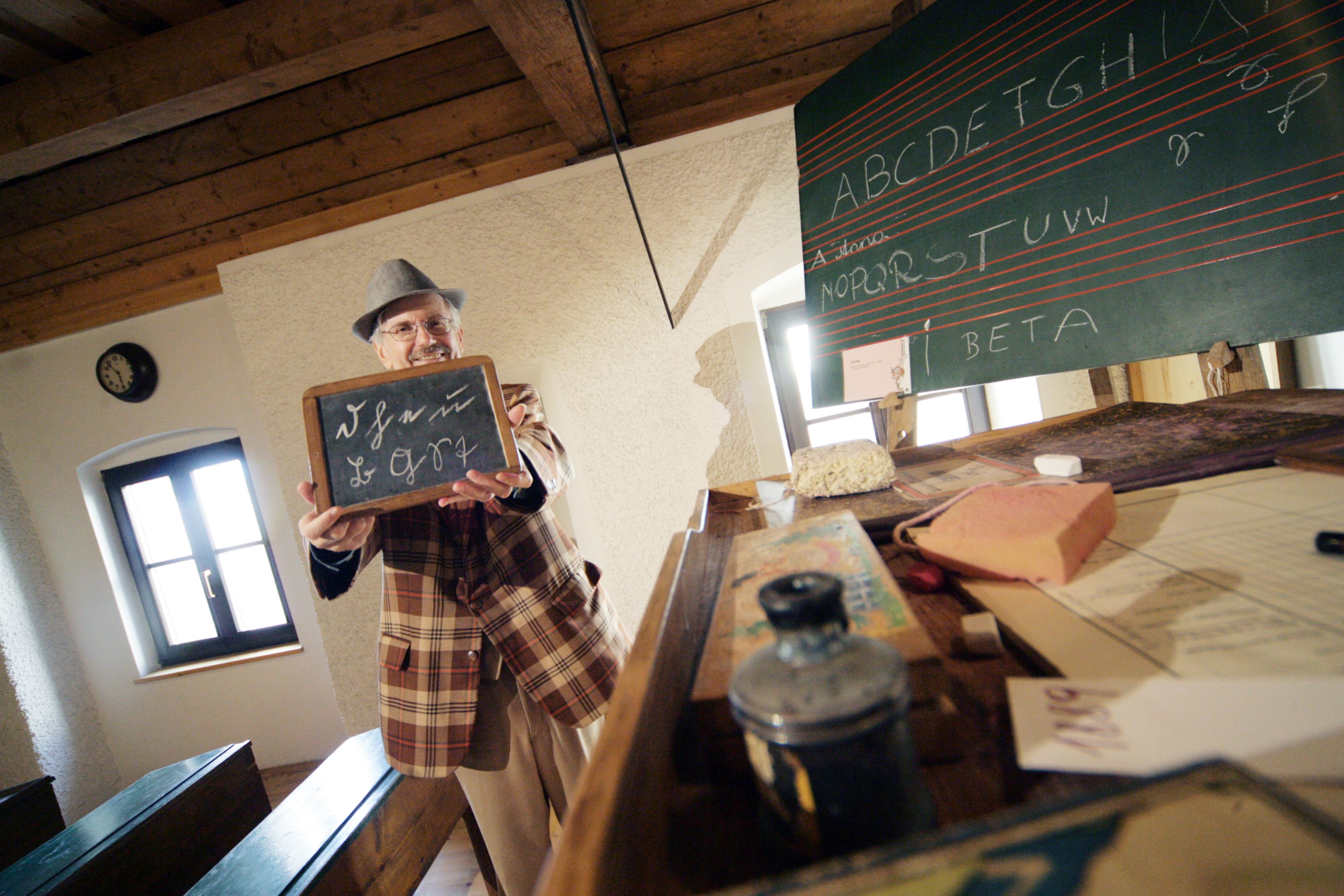Ein Mann in einem karierten Anzug hält eine Schiefertafel in einem historischen Klassenzimmer.