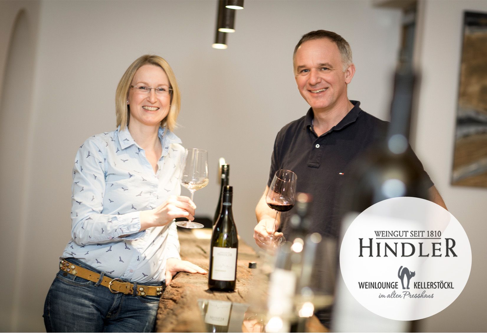 Ein Winzerpaar steht lächelnd mit Weingläsern in der Hand an einem Tisch im Weingut Hindler.