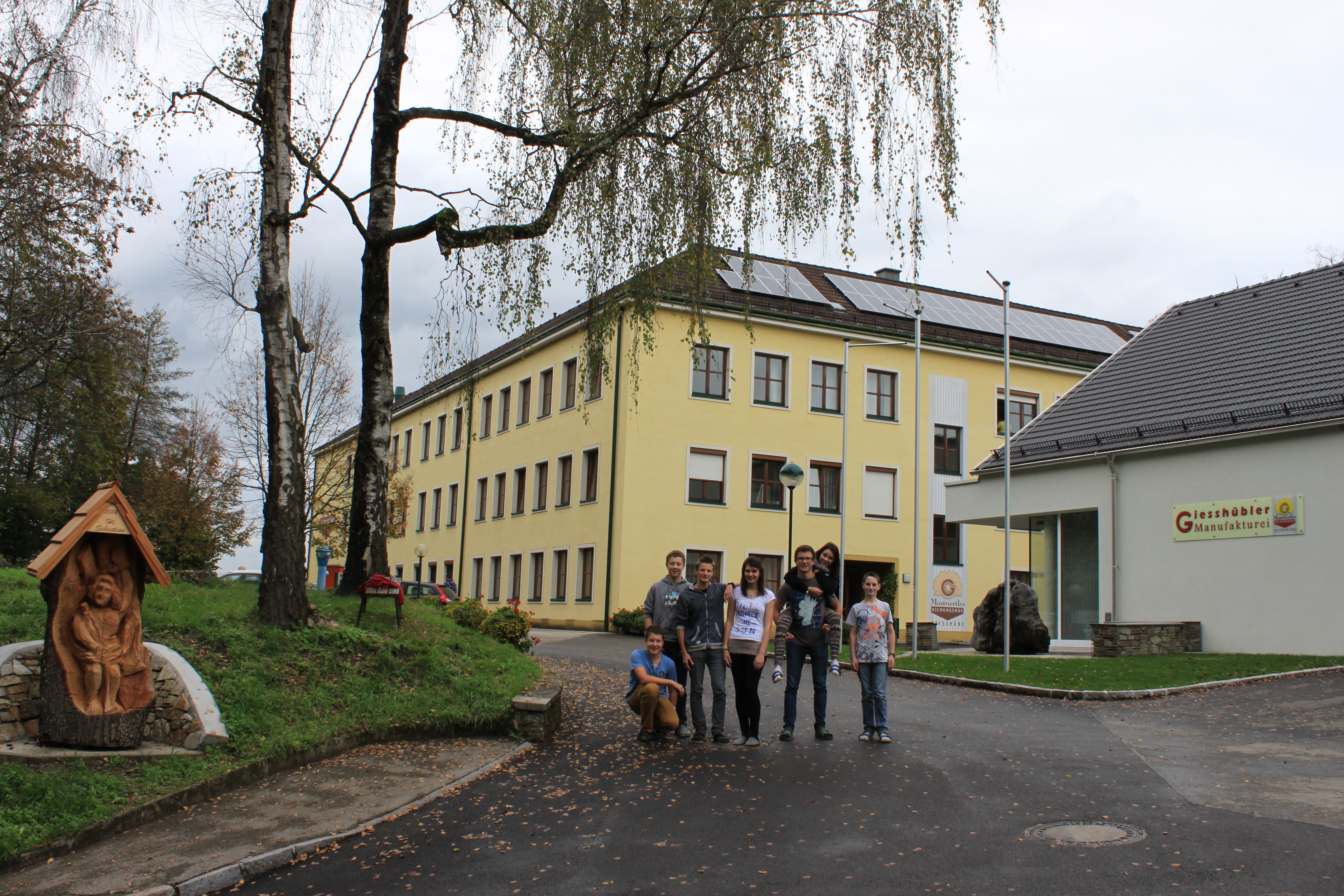 Gruppe von Menschen vor einem gelben Schulgebäude mit Solarpanelen auf dem Dach.