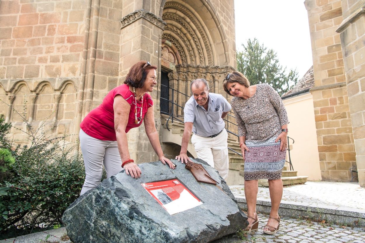 Drei Personen betrachten eine Gedenktafel auf einem Stein vor einem historischen Gebäude.