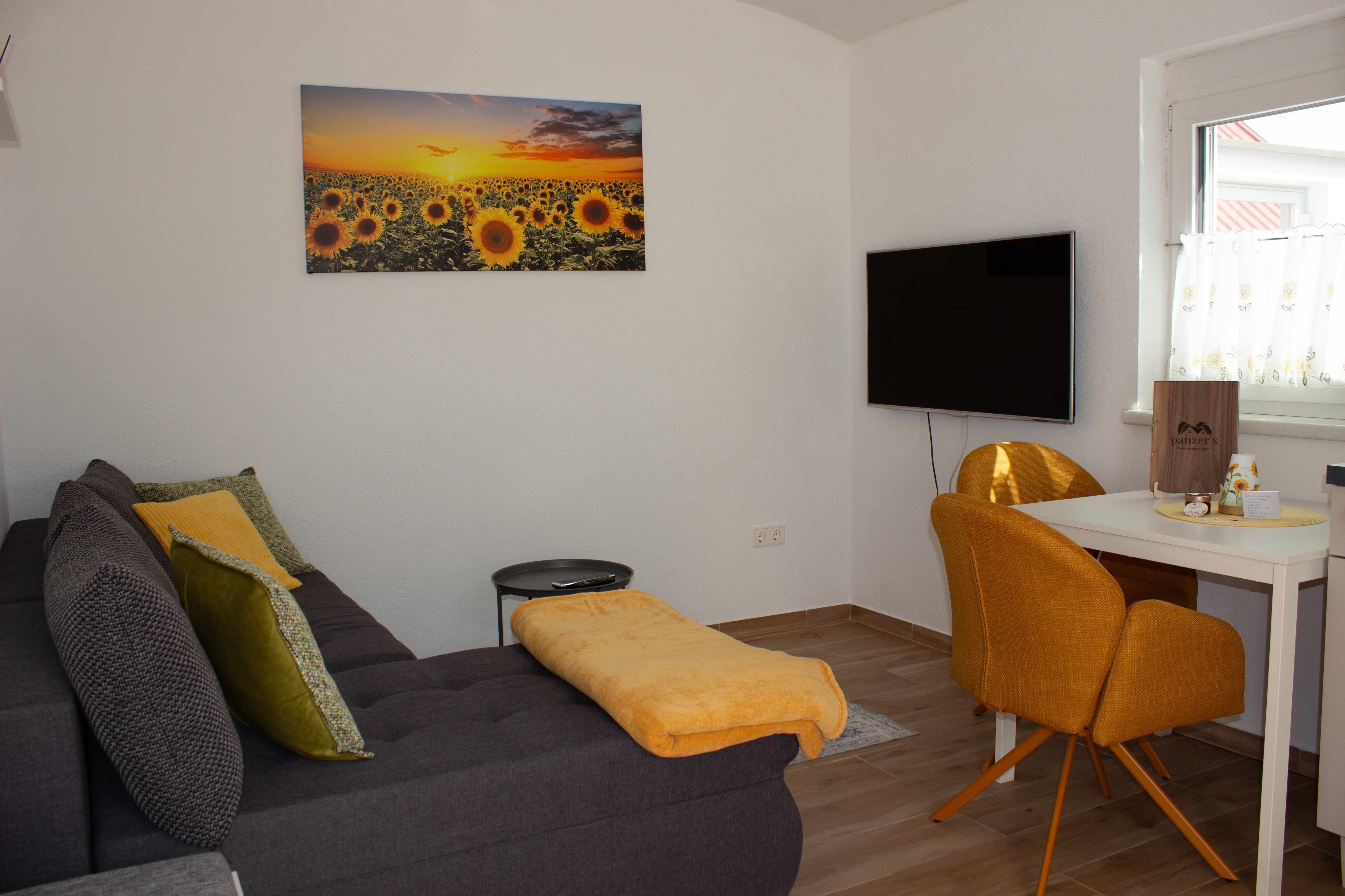 Wohnzimmer einer Ferienwohnung mit grauem Sofa, gelben Kissen, einem Bild von Sonnenblumen und einem Fernseher an der Wand.