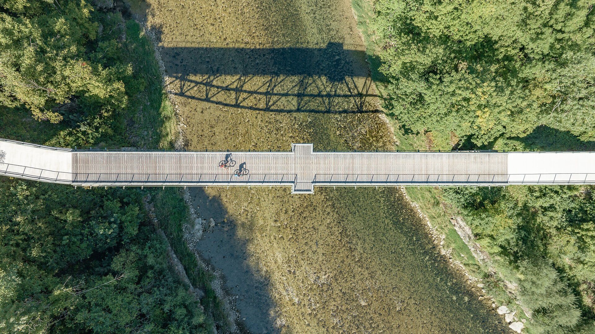 Eine malerische Brücke erstreckt sich über das glitzernde Wasser, während Radfahrer die frische Bergluft genießen. Umgeben von üppigem Grün und majestätischen Bäumen, lädt die Landschaft zu unvergesslichen Erlebnissen in der Natur ein.