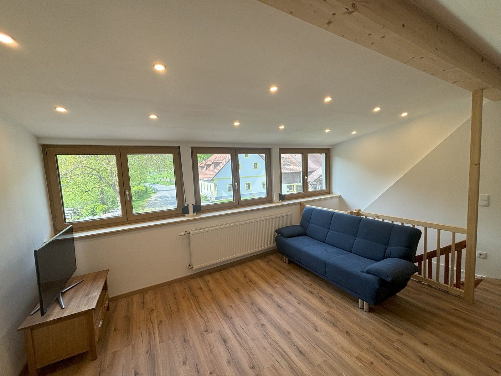 Wohnzimmer mit blauer Couch, Holzboden und großen Fenstern mit Blick nach draußen.