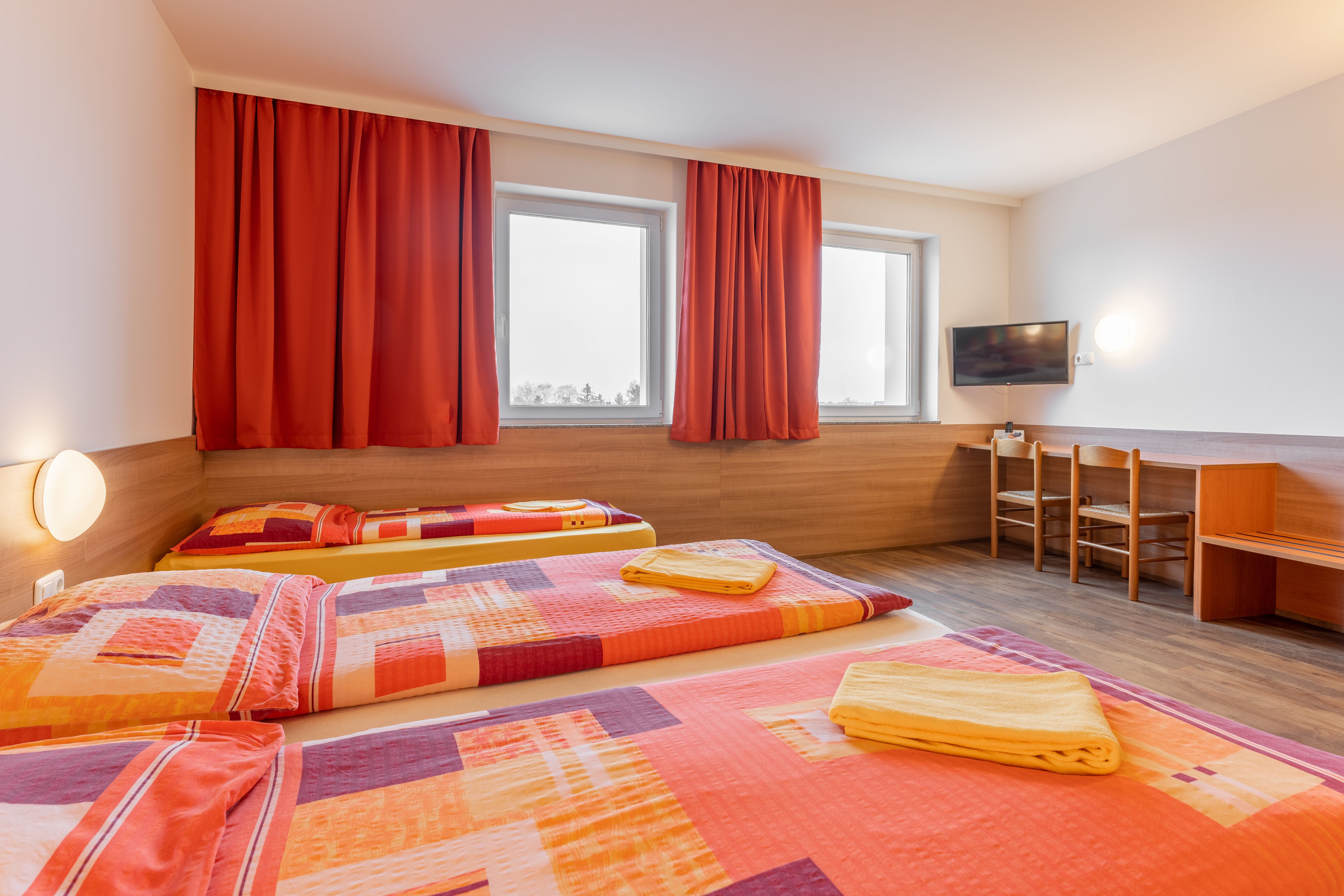 Hotelzimmer mit drei Betten, roten Vorhängen und einem Fernseher an der Wand.