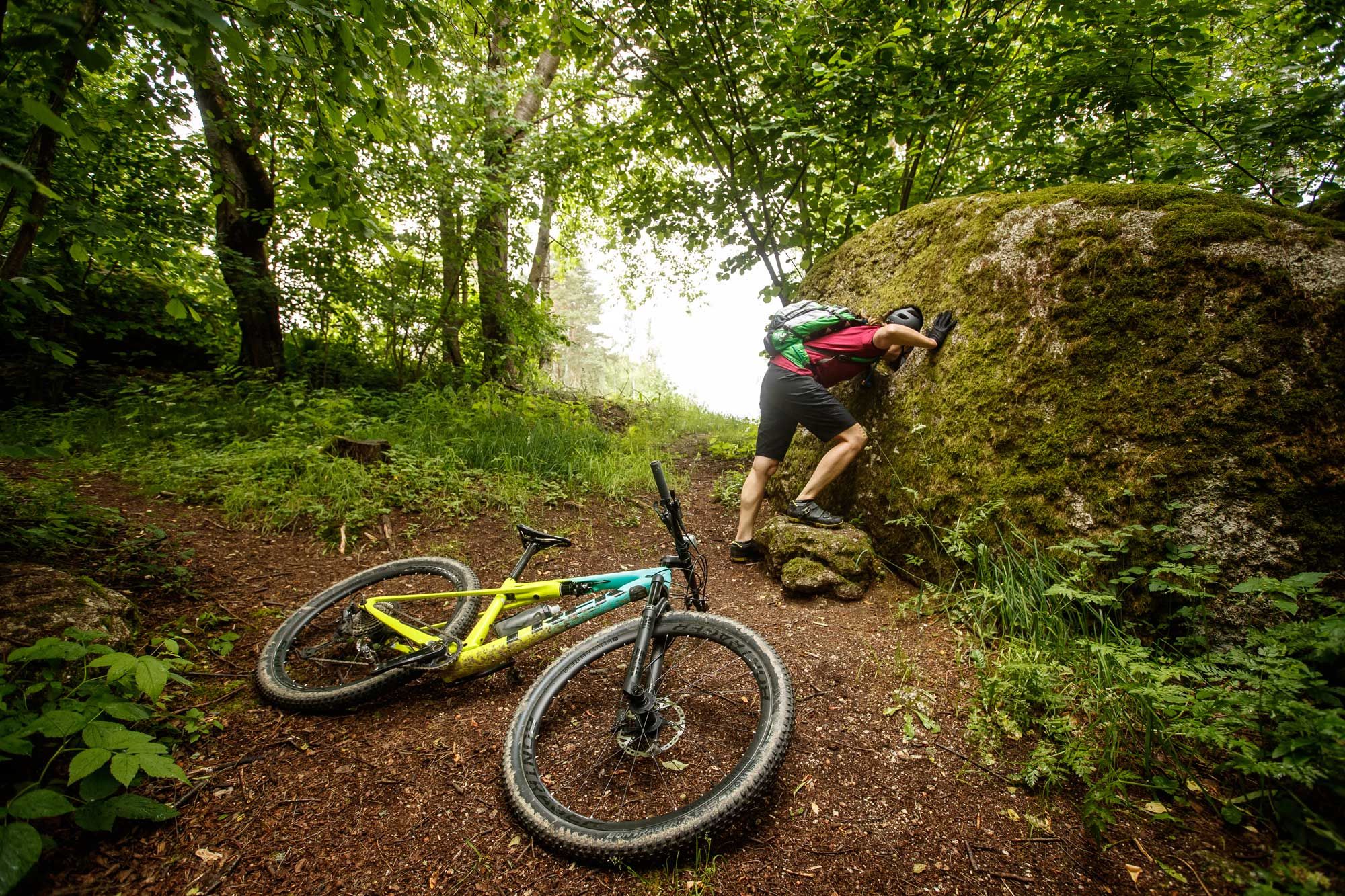 Ein Mountainbiker lehnt an einem großen, moosbedeckten Felsen im Wald, während sein Fahrrad auf dem Boden liegt.