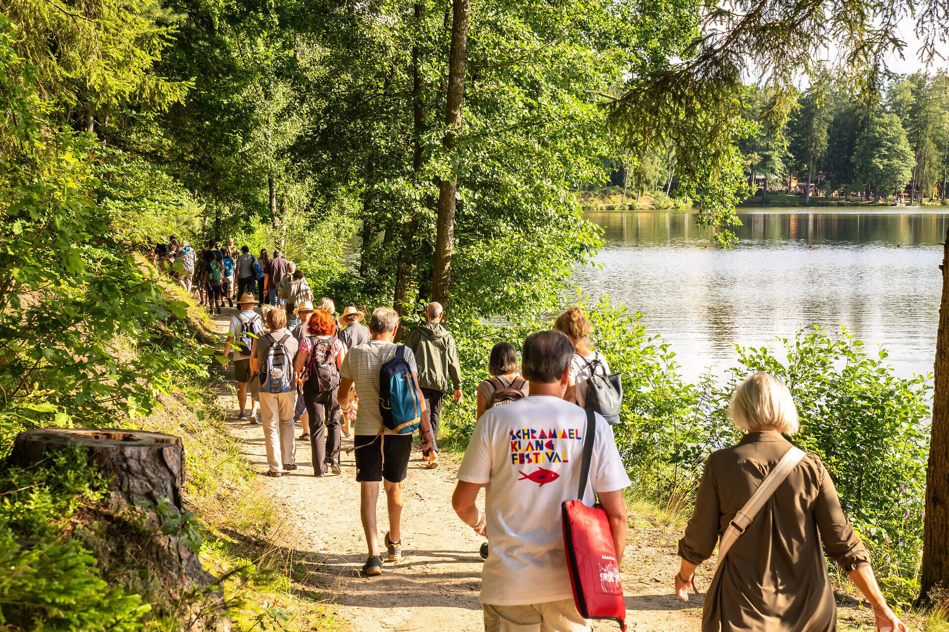 Eine Gruppe von Menschen wandert entlang eines malerischen Weges, umgeben von üppigem Grün und dem sanften Plätschern des Wassers. Die Atmosphäre ist lebhaft und fröhlich, während die Teilnehmer des Schrammel.Klang.Festivals die Schönheit der Natur und die Freude an der Musik genießen.