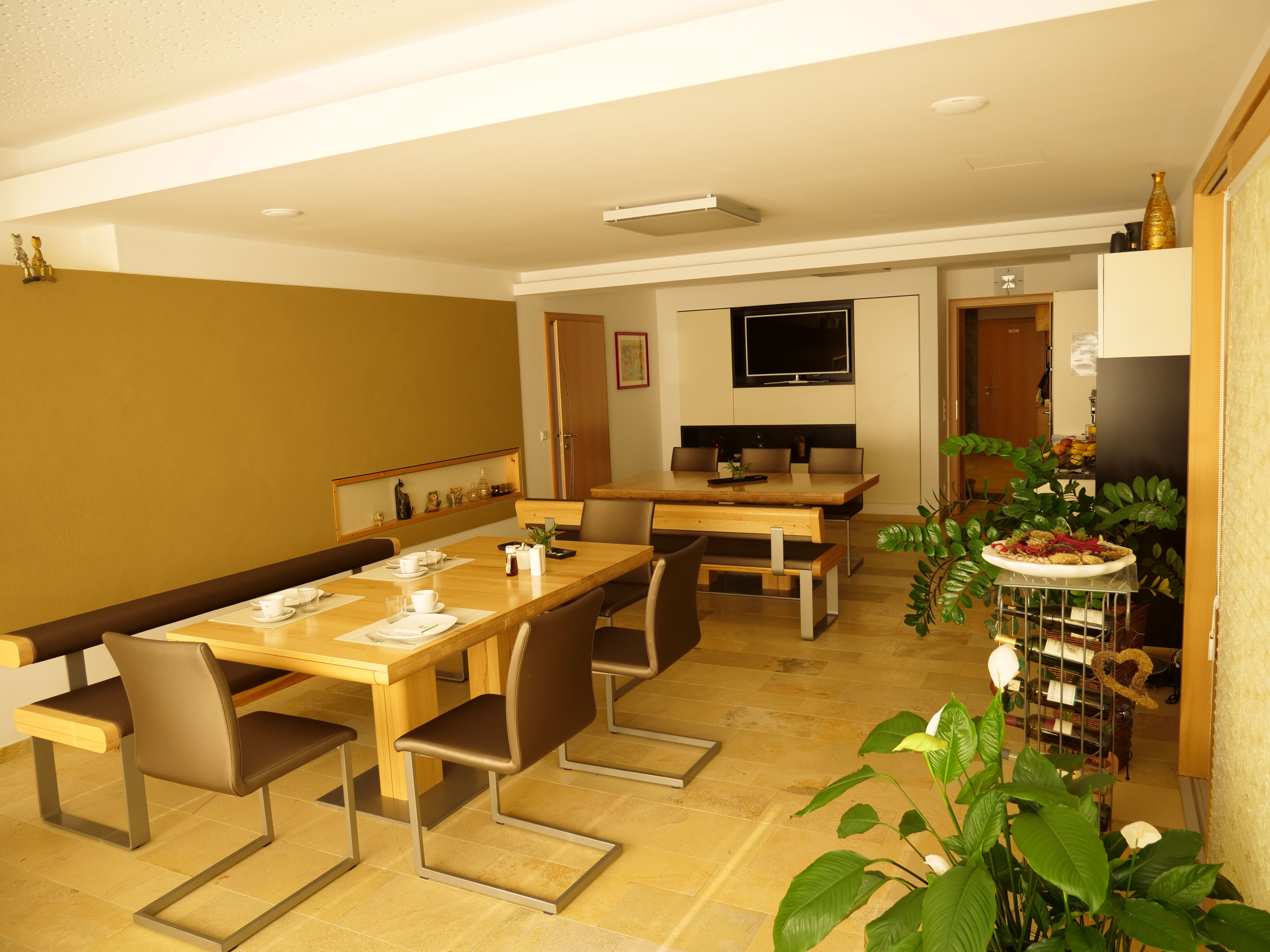 Ein Frühstücksraum mit Holztischen, Stühlen und Pflanzen, dekoriert mit Geschirr und einem Fernseher an der Wand.