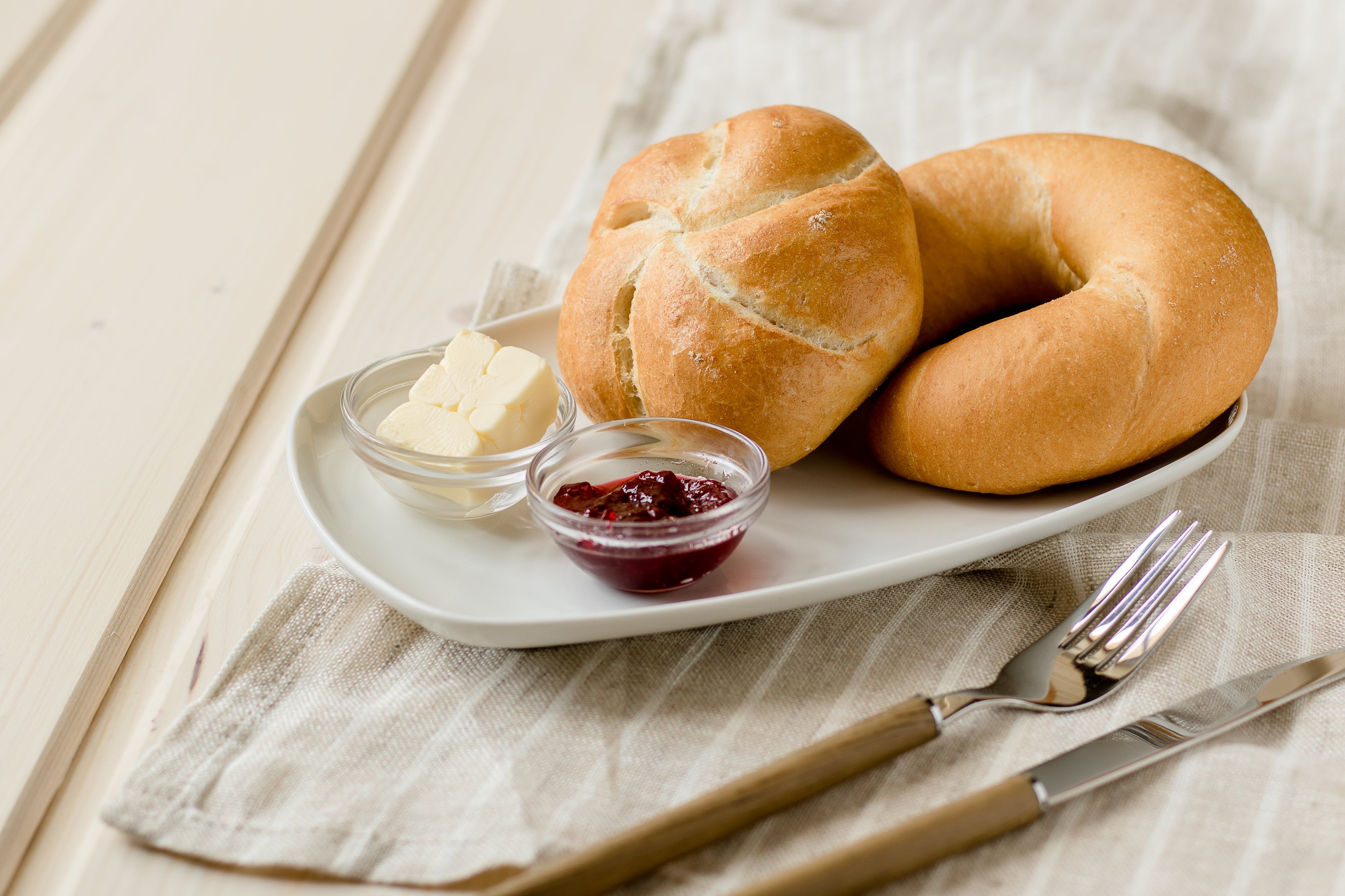 Ein Teller mit einer Semmel und einem Kipferl, Butter und Marmelade auf einem Tisch mit Besteck.