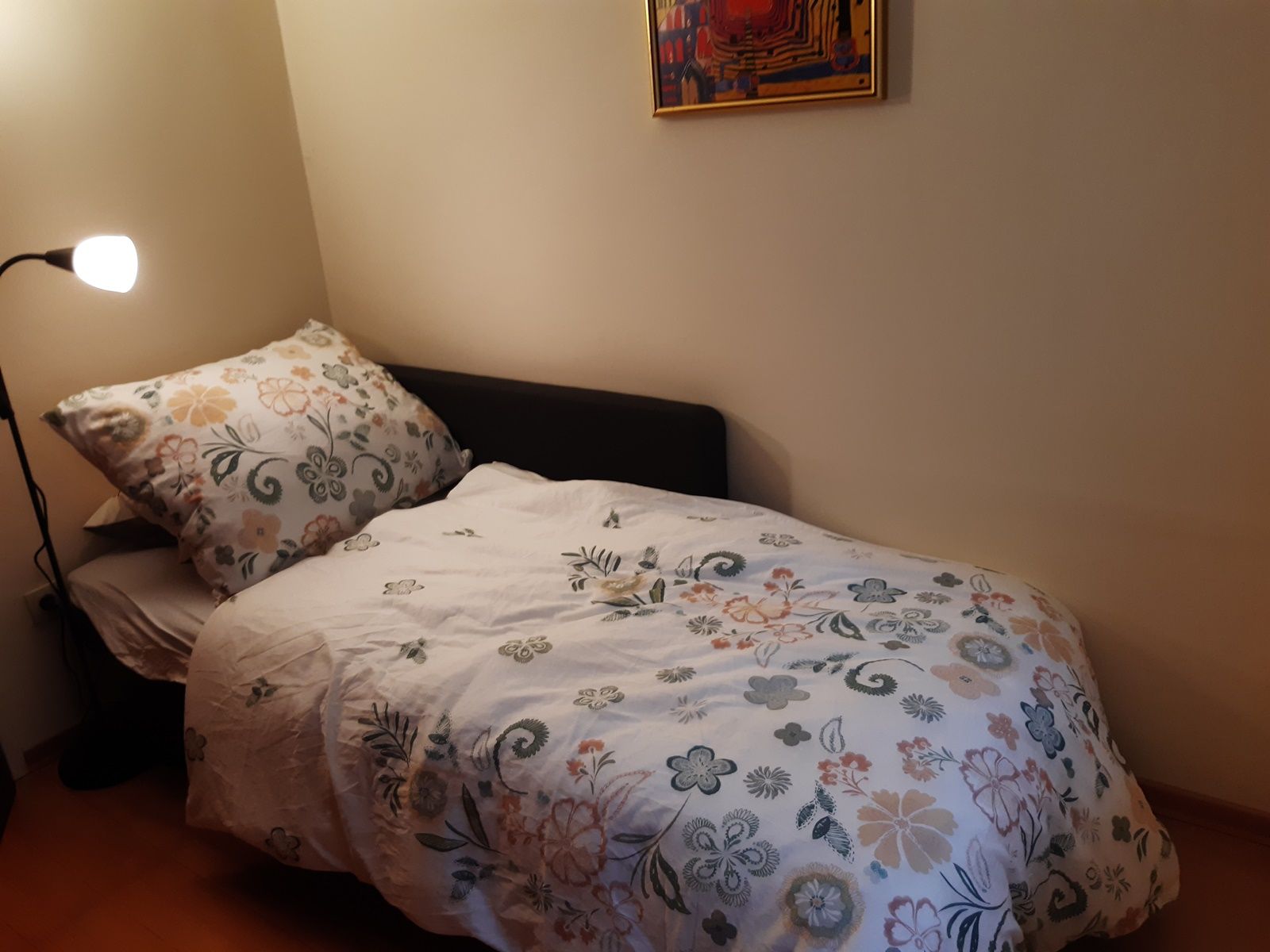 Ein kleines Zimmer mit einem Einzelbett, das mit einer geblümten Bettdecke bezogen ist. Eine Stehlampe steht daneben, und ein buntes Bild hängt an der Wand.