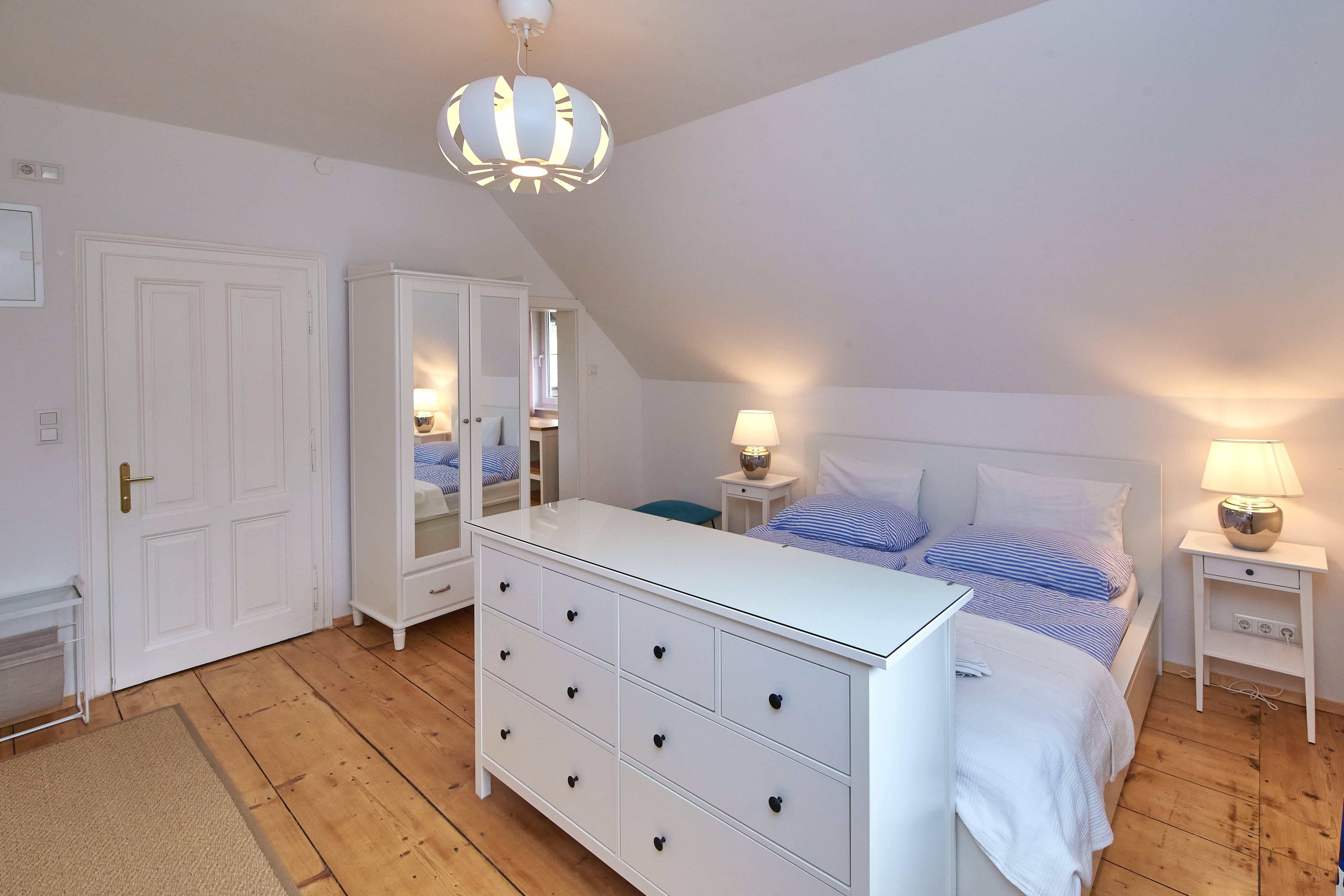 Helles Schlafzimmer mit Doppelbett, weißem Kleiderschrank und Kommode, Holzboden und moderner Deckenlampe.